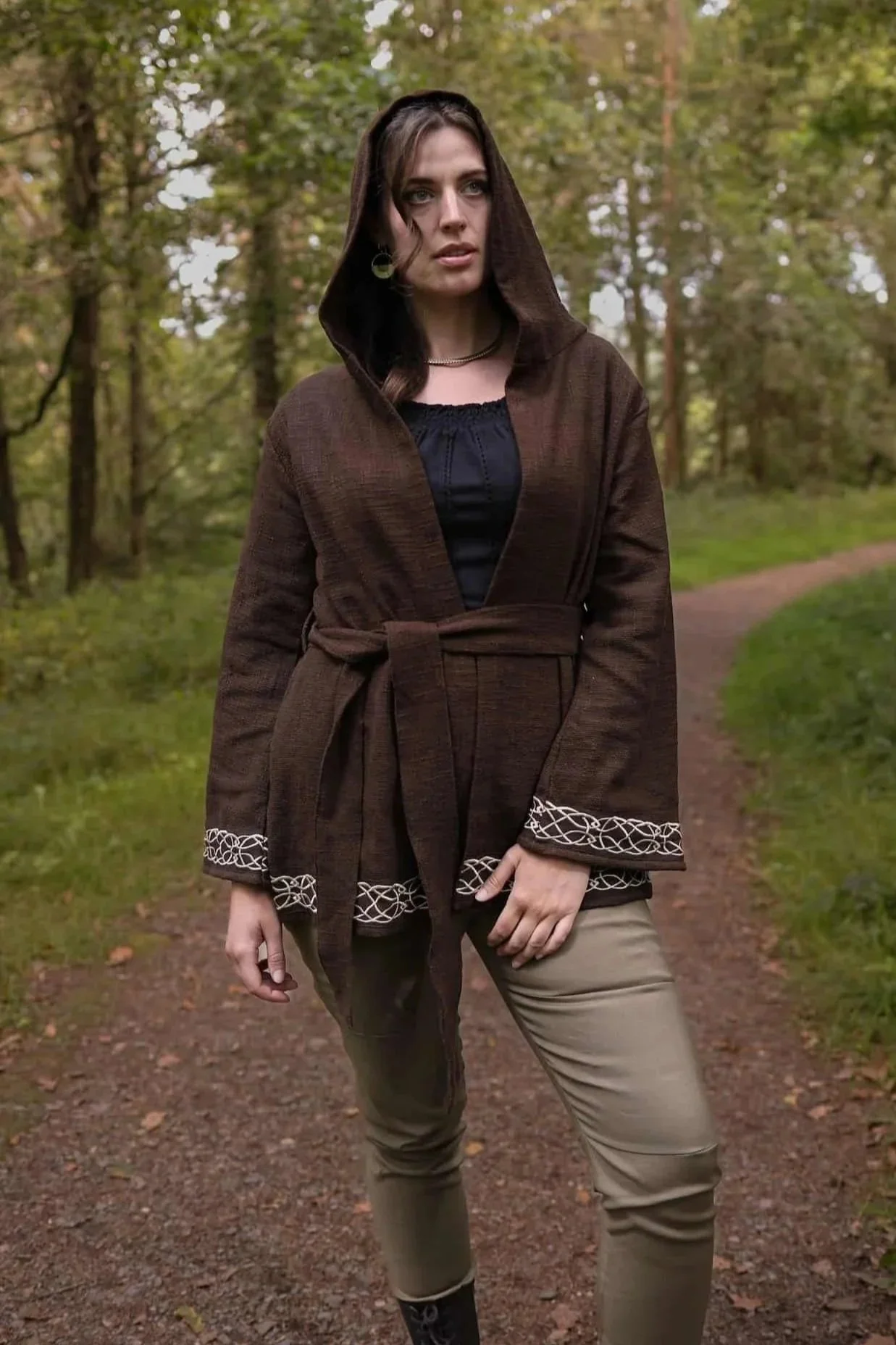 Woman wearing Celtic Fusions handwoven cotton hooded wrap cardigan with Celtic knotwork embroidery on cuffs and hem; belted at waist. Folklore cardigan photographed in a woodland setting.