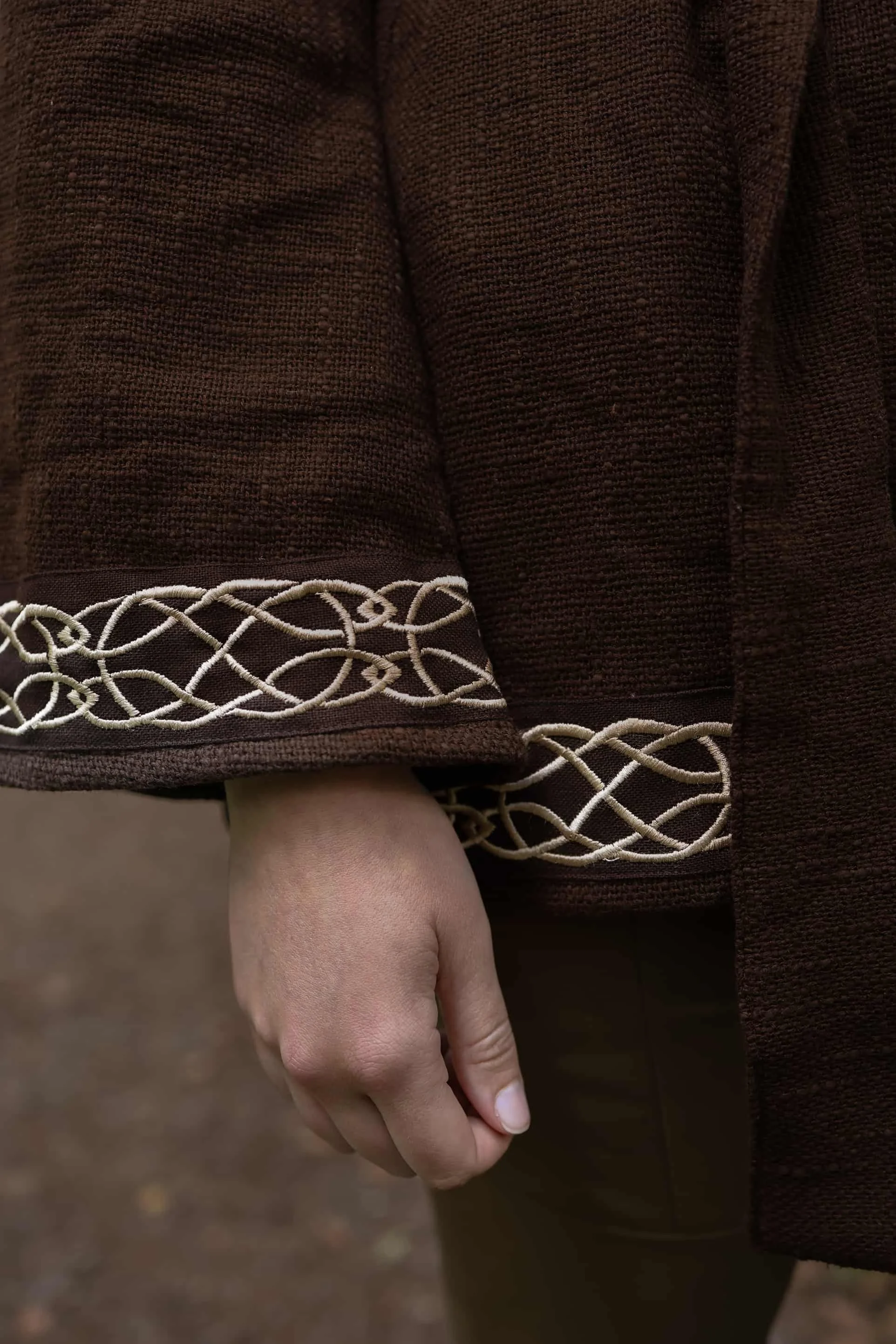 Woman wearing Celtic clothing handwoven wrap cardigan with Celtic knot embroidery on cuffs and hem; belted at waist. Folklore cardigan photographed in a woodland setting.