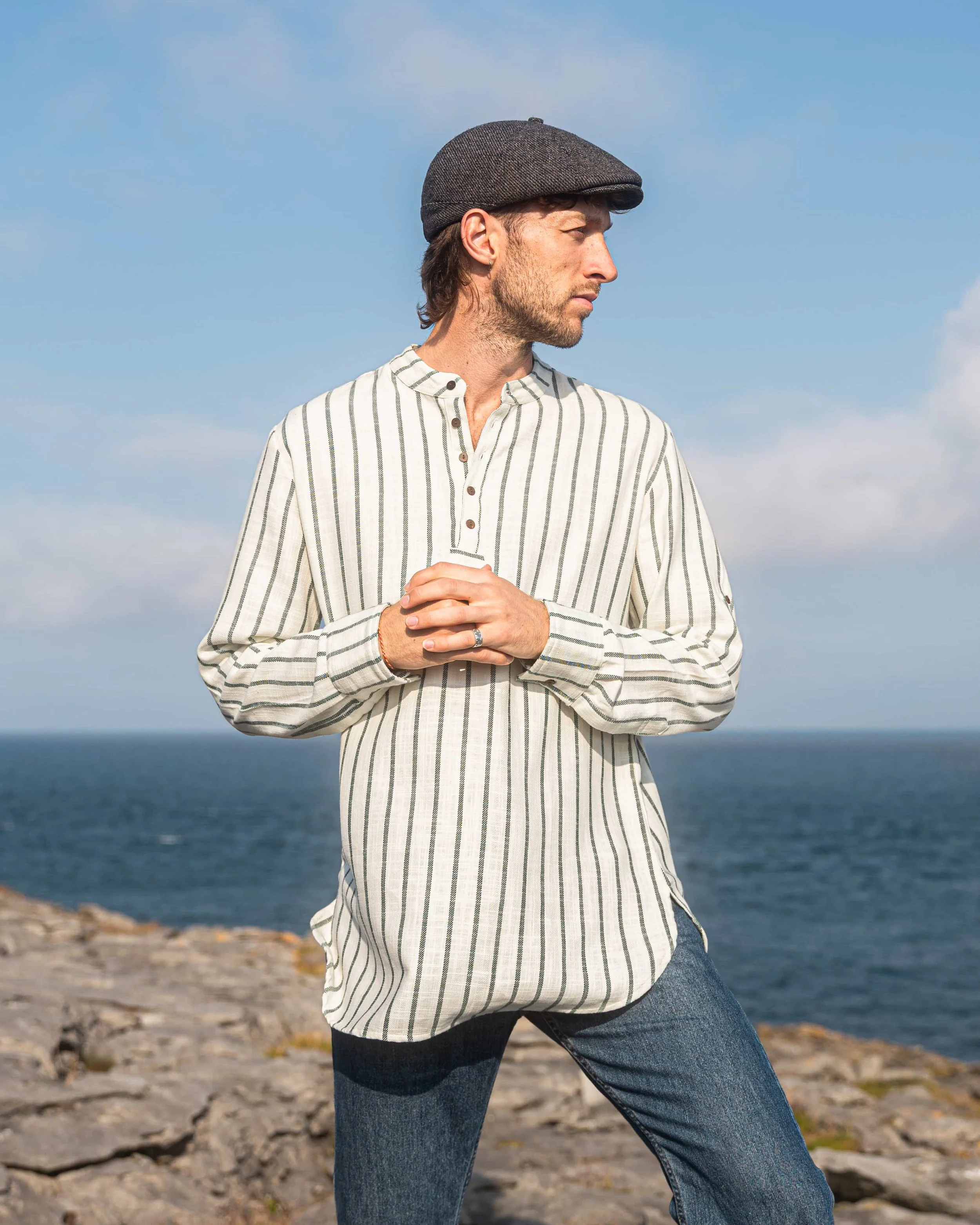 Model wearing a forest green and cream striped collarless shirt in 100% cotton, with coconut buttons and rolled sleeves, pictured by the Atlantic coast.