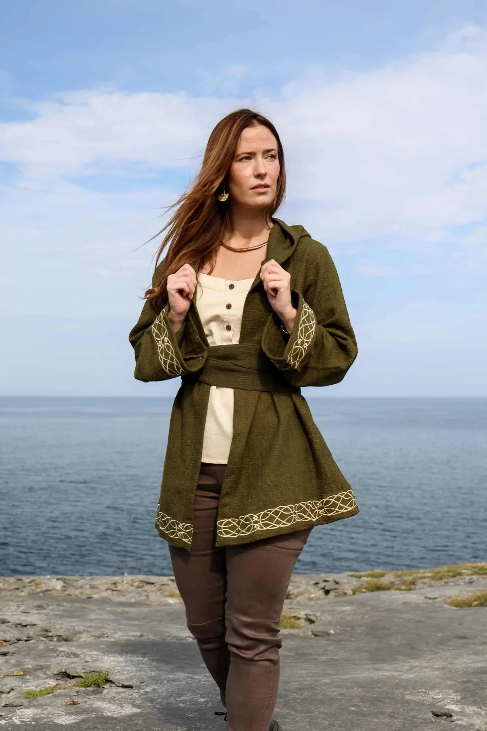 Woman wearing Celtic clothing handwoven wrap cardigan with Celtic knot embroidery on cuffs and hem; belted at waist. Folklore cardigan photographed in a woodland setting.