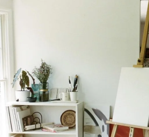 A cozy corner of a room with a white bookshelf holding books, plants, and a vase, next to an easel with a blank canvas.