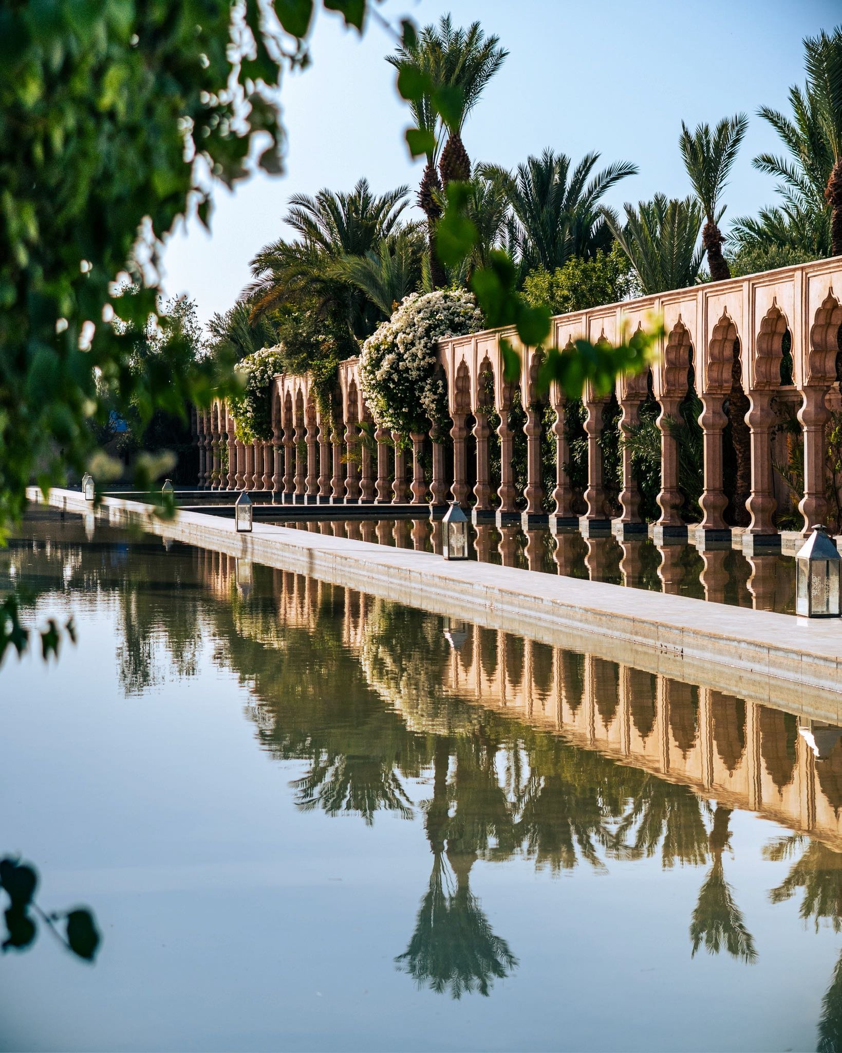 A tranquil canal with reflections of palm trees and a decorative wall topped with flowering bushes, set against a clear blue sky.