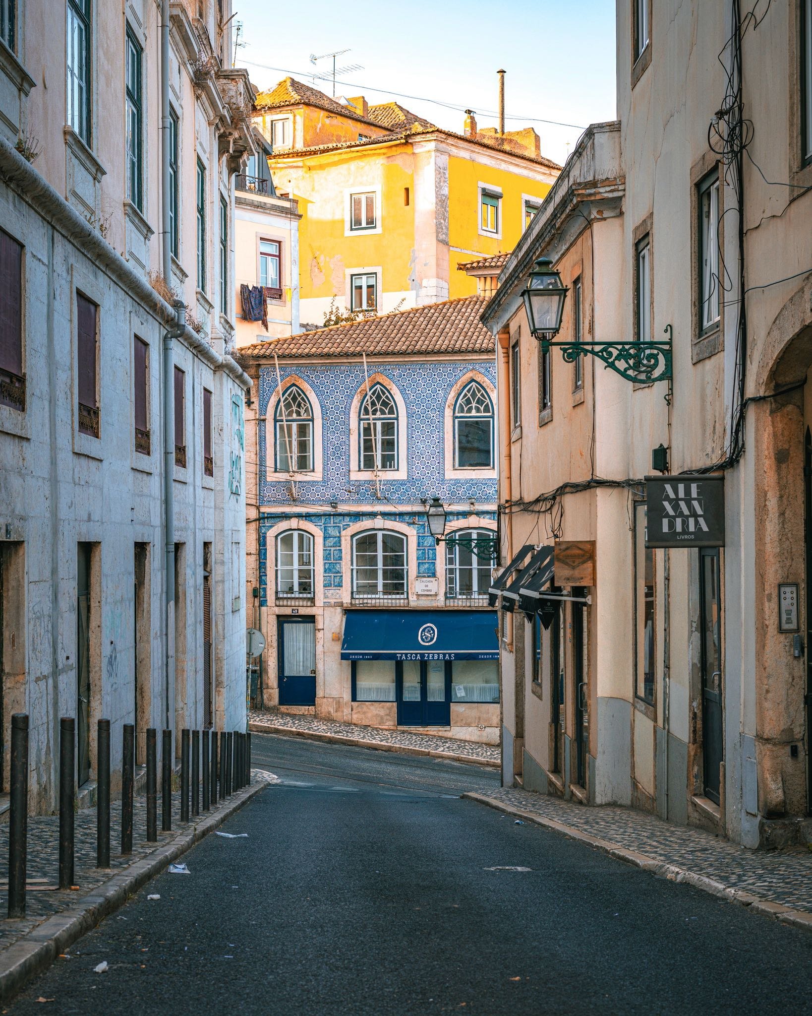A narrow, winding street in a European city with colorful buildings, one featuring blue tiles and arched windows, and a yellow building in the background. The street has a cobblestone sidewalk and black bollards on the side.