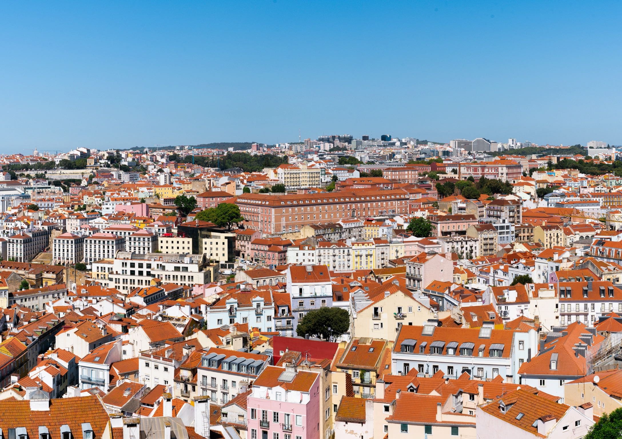 Panoramic view of Lisbon, Portugal, featuring a cityscape with red-tiled roofs, colorful buildings, and some trees under a clear blue sky.
