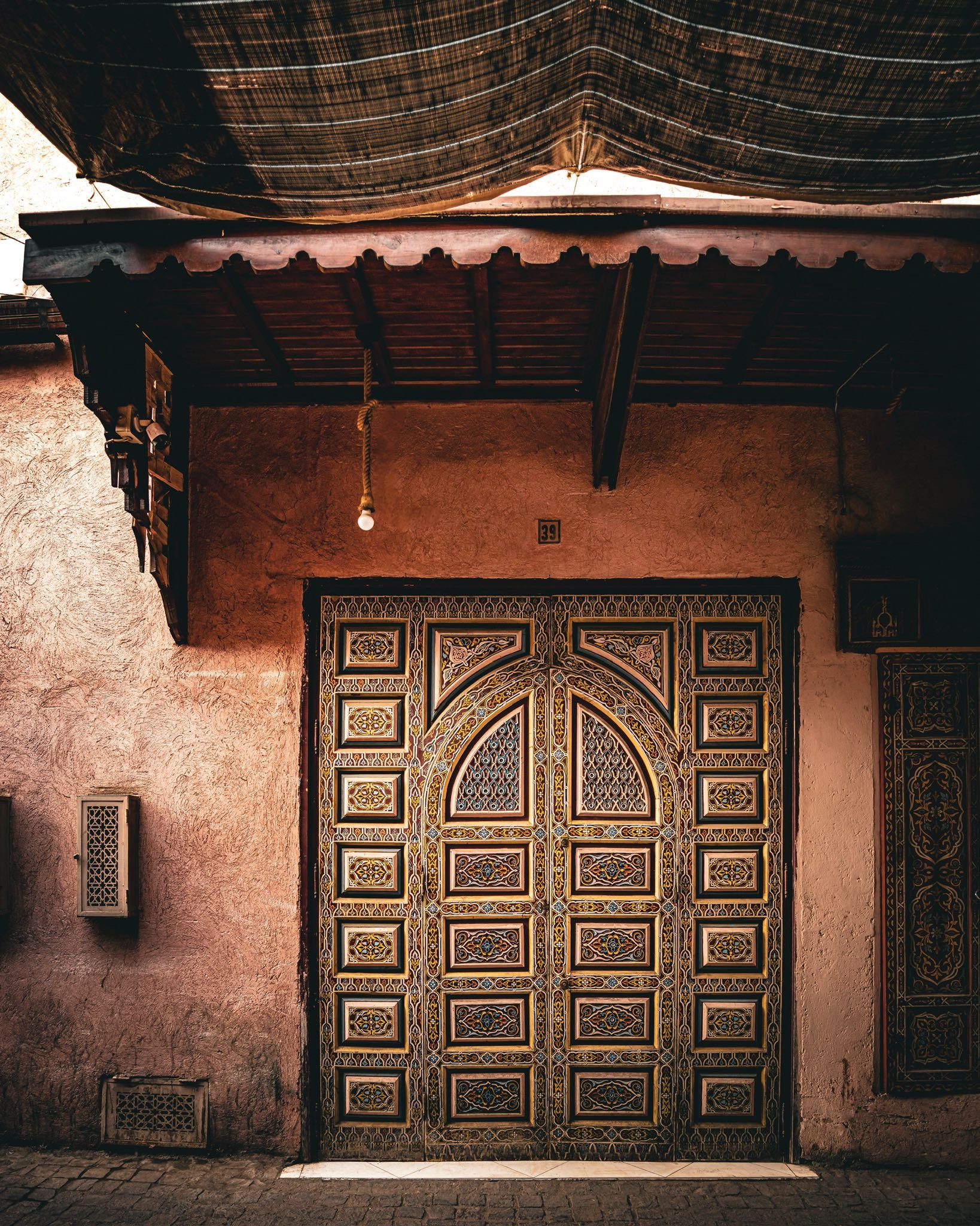 Decorative wooden door with intricate gold, black, and blue patterns on a pinkish textured wall, with a small window to the right and a small vent beneath, under a wooden roof with a hanging light bulb.
