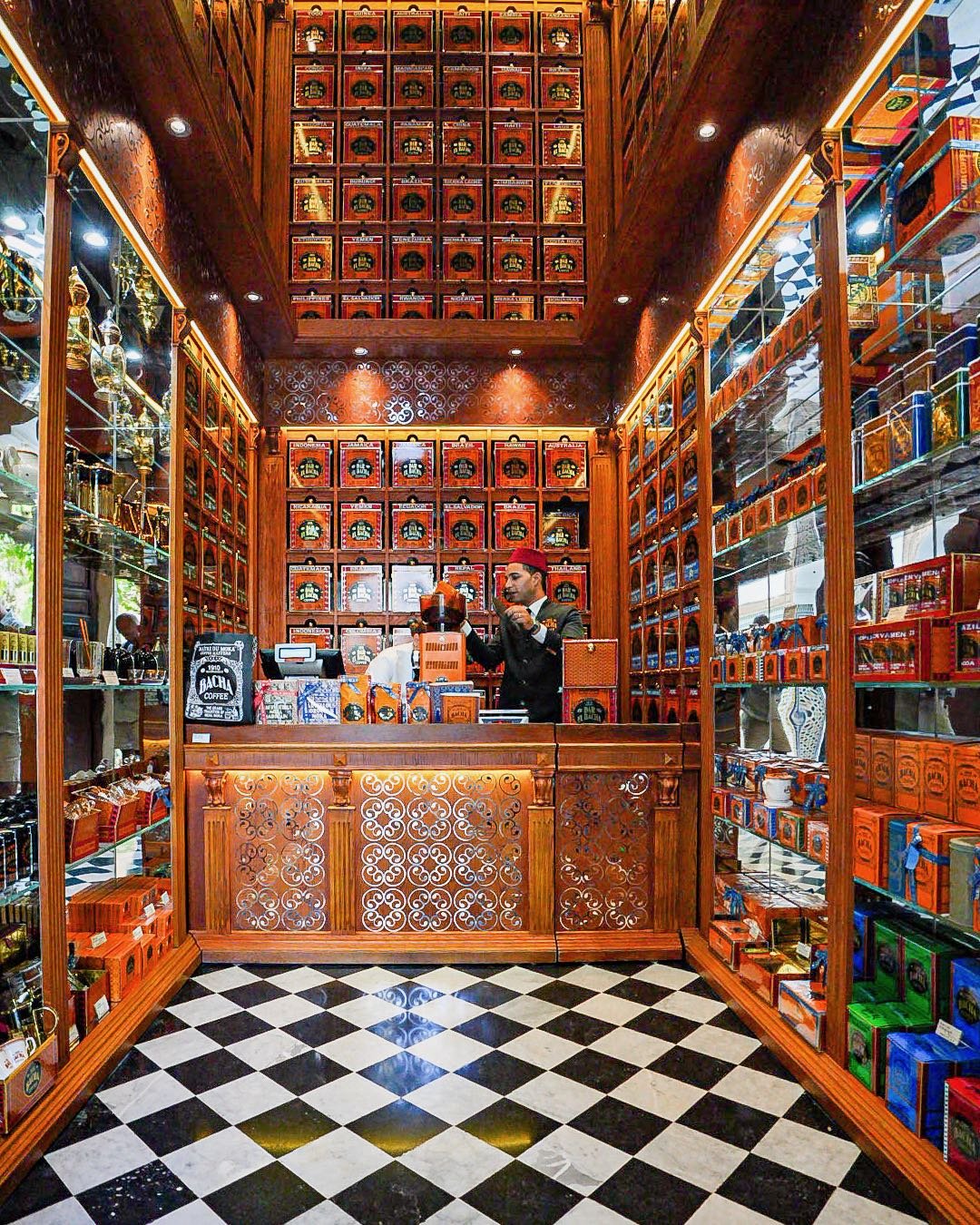 A coffee shop with dark wooden fixtures, shelves filled with orange and multicolored boxes, and a man in a dark uniform and red hat preparing coffee behind the counter. The floor features a black and white checkered tile pattern.