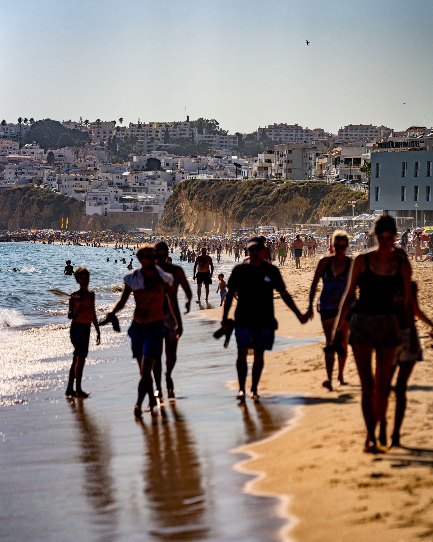 People walking along the sandy beach near the sea with cliffs and white buildings in the background on a sunny day.