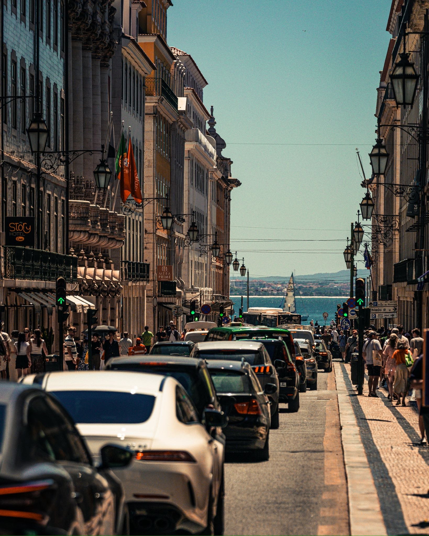 A busy city street lined with historic buildings, with cars parked on one side and pedestrians walking on the sidewalk, leading to a body of water with a sailboat in the background under a clear sky.