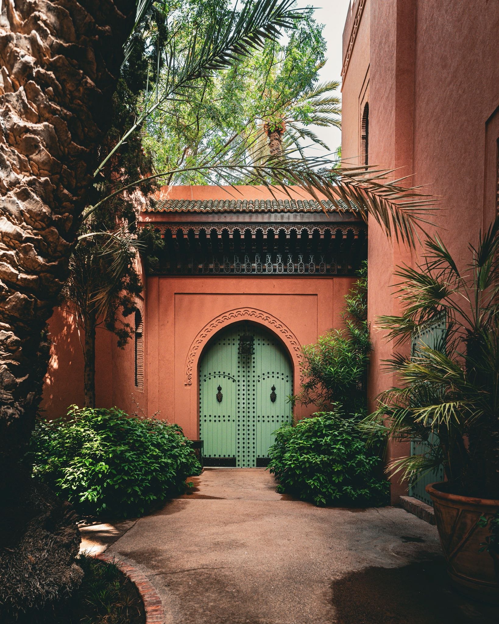A pink stucco building with a green arched door, surrounded by lush green plants and tall trees, in a courtyard with a concrete walkway.