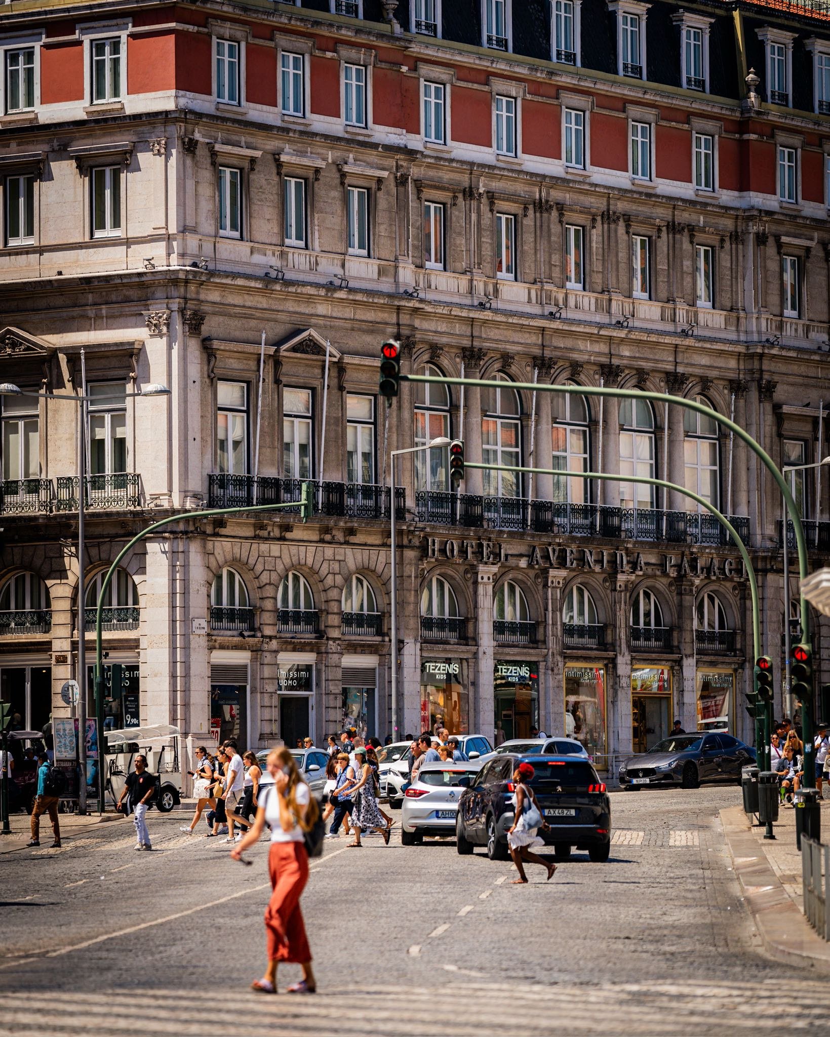 Street scene with people crossing at a crosswalk, cars waiting, and a historic hotel building in the background with signs for shops on the ground floor.