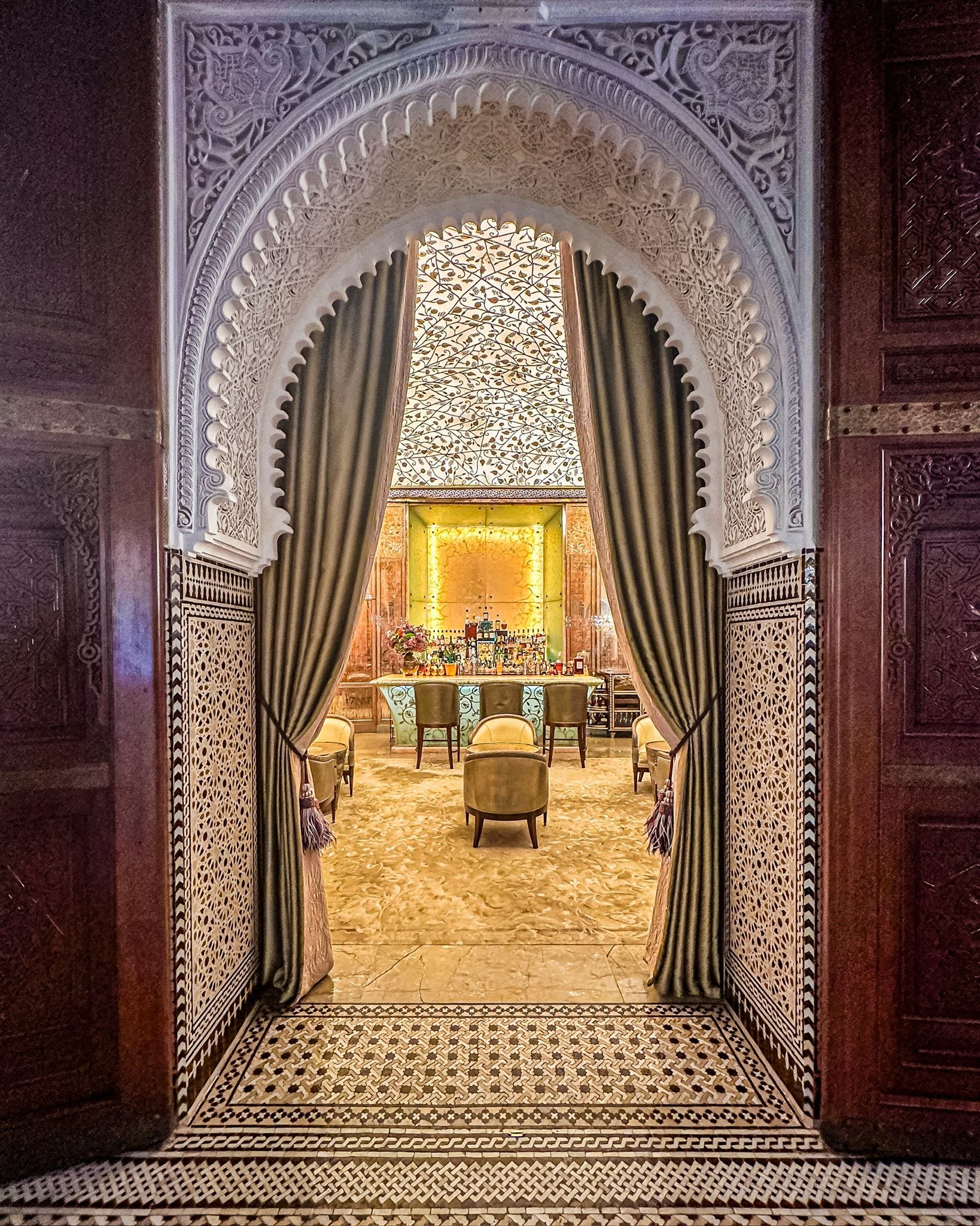 Elegant indoor lounge with ornate Moroccan-style archway, patterned ceiling, velvet curtains pulled to the sides, plush chairs, and a bar area in the background with warm lighting.