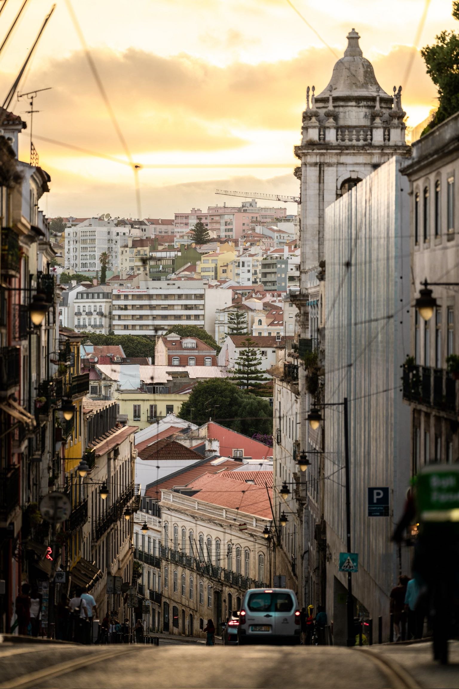 A city street at sunset with buildings on both sides, a car driving down the hill, and a view of colorful rooftops and high-rise buildings in the distance under a cloudy sky.