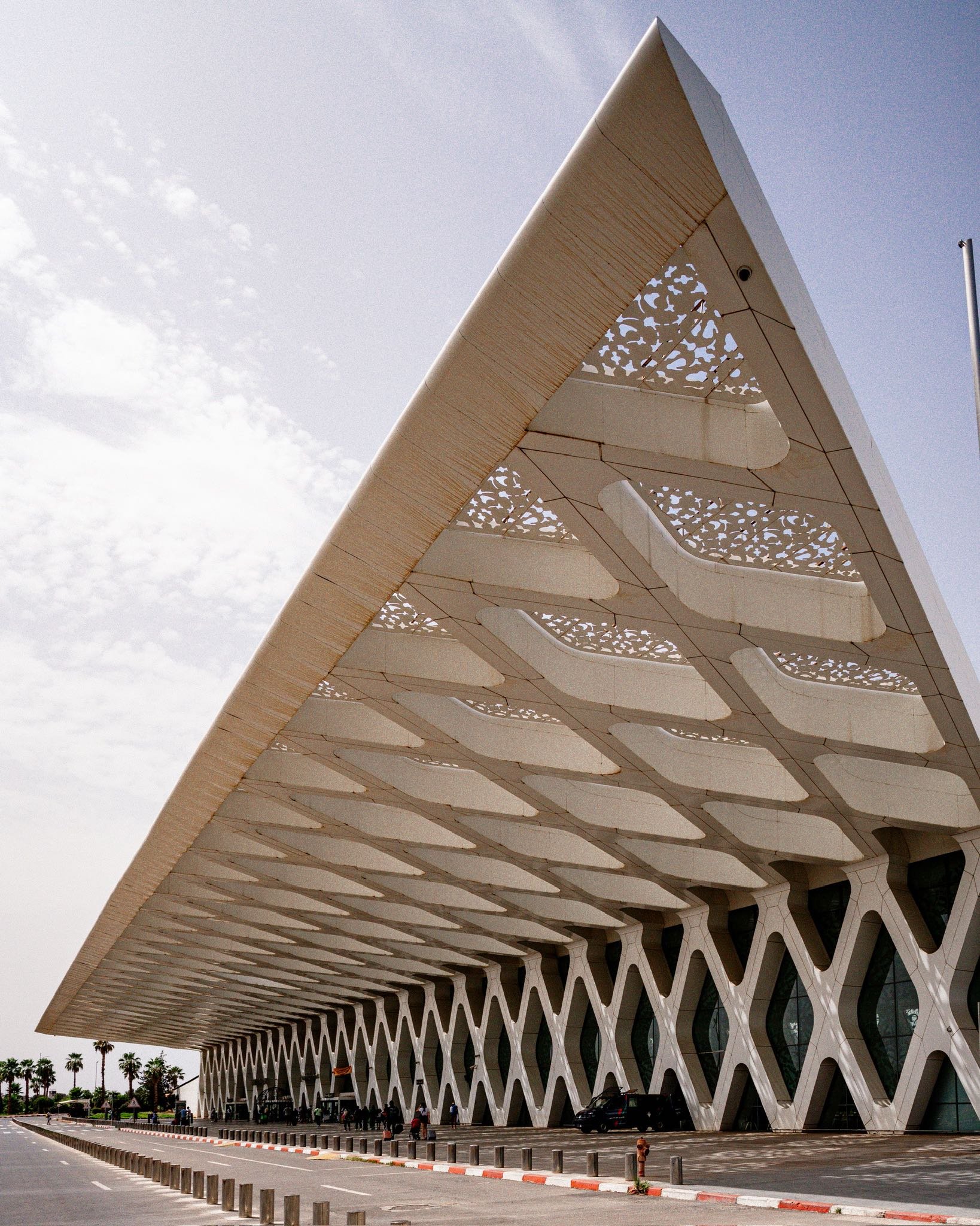 Modern building with a large, overhanging roof and a facade featuring a diamond pattern, located along a street with a few cars and palm trees in the background.