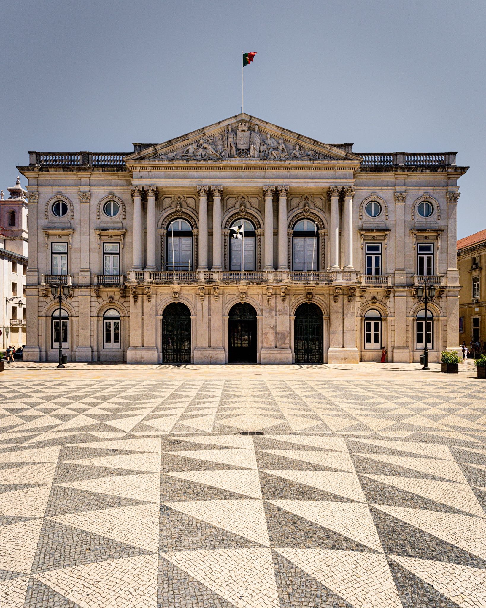 Historical building with classical architecture, columns, and a flag on top, in a city square with patterned pavement.