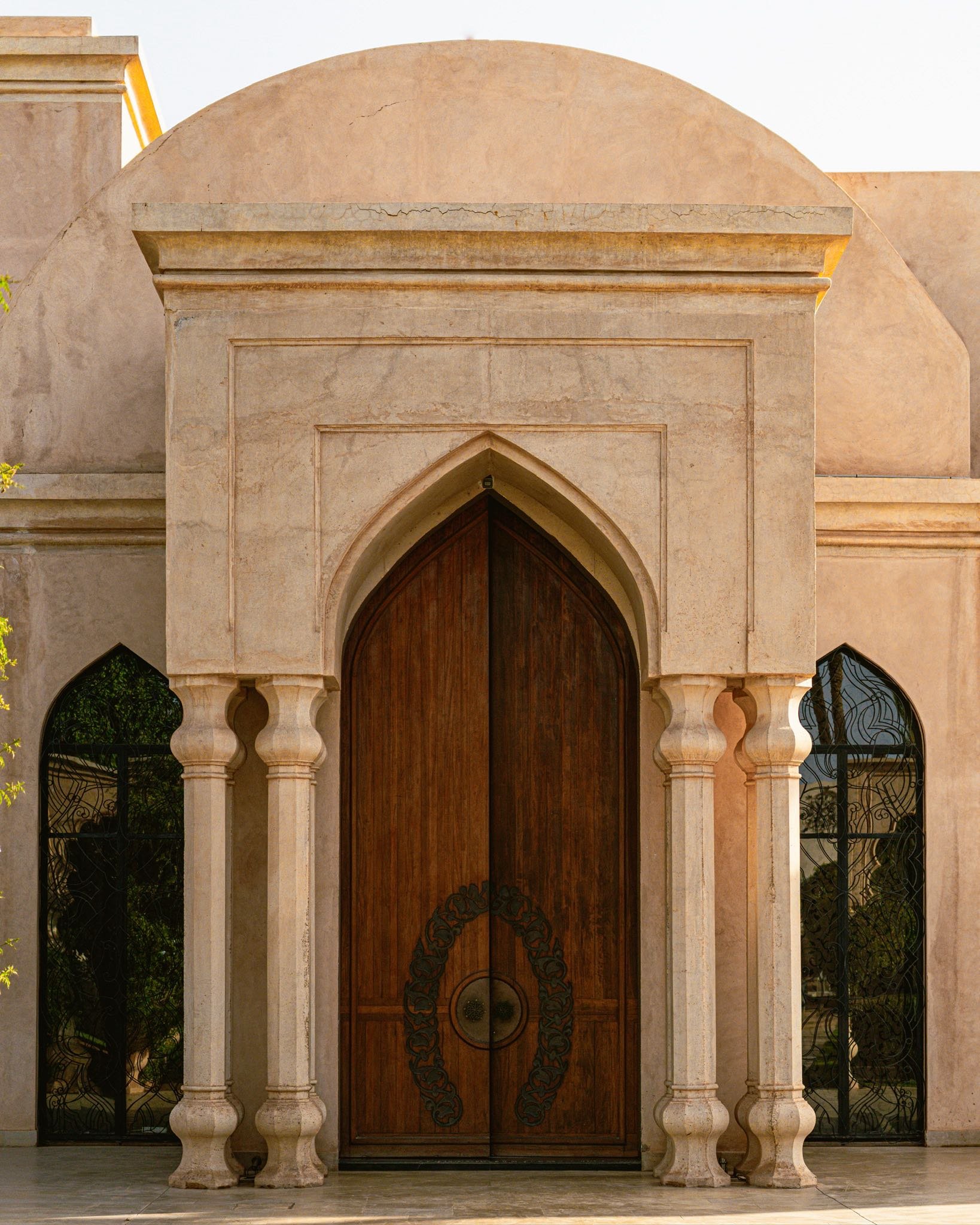 A grand wooden door with pointed arch design framed by stone columns, with two arched glass windows on either side reflecting trees.