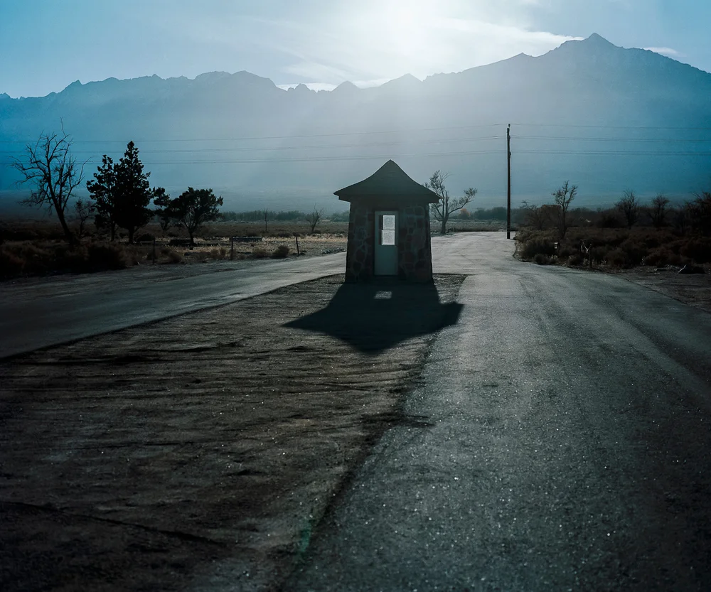 Color photograph of a small stone guard house on an empty road at dusk, with mountains behind it.