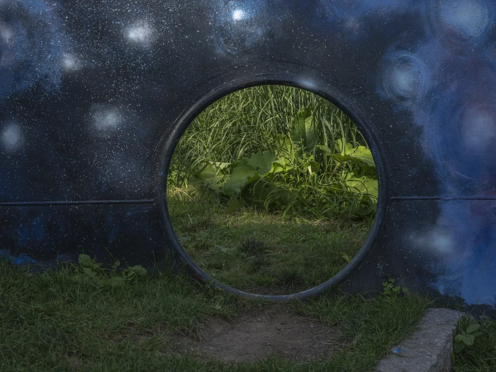 Color photograph of a round opening in a dark blue mural dotted with white stars, framing a bright green garden beyond.