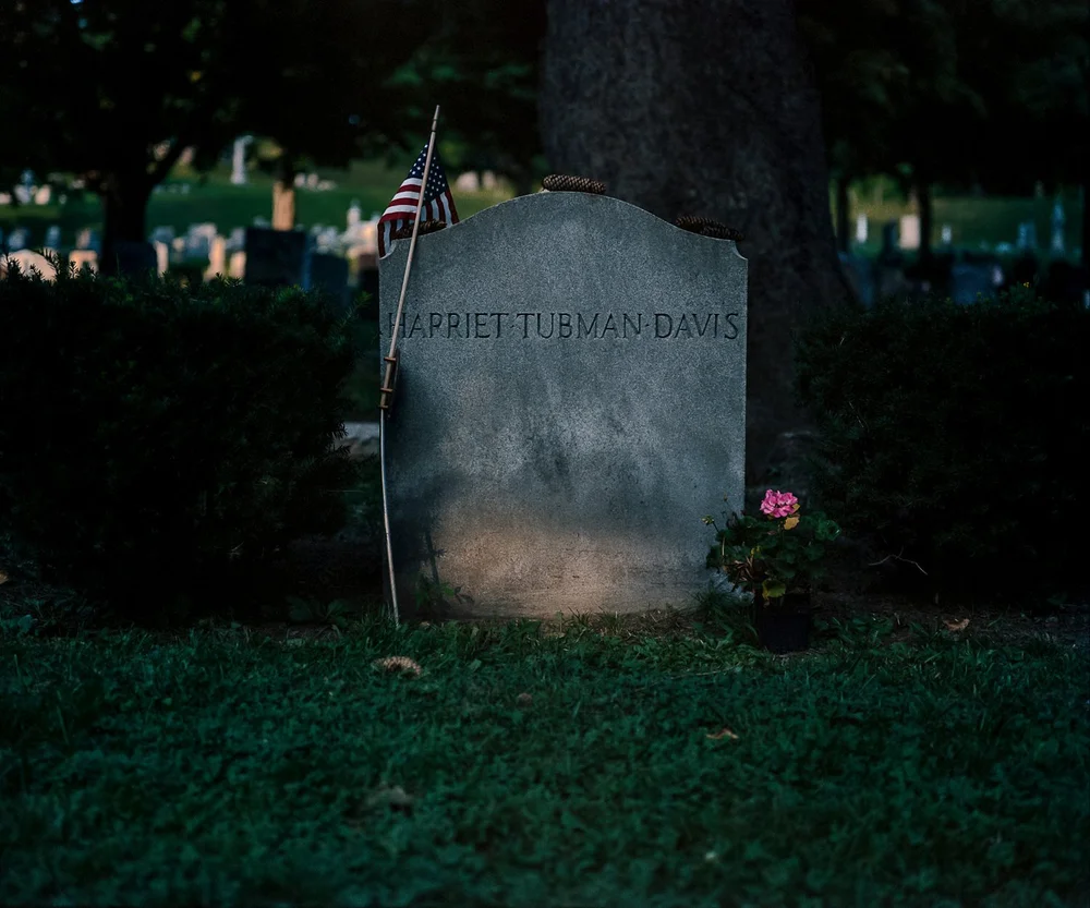 Color photograph of a weathered gray gravestone engraved with the name Harriet Tubman Davis, with a small flag and pink flowers at its base, under a large tree.