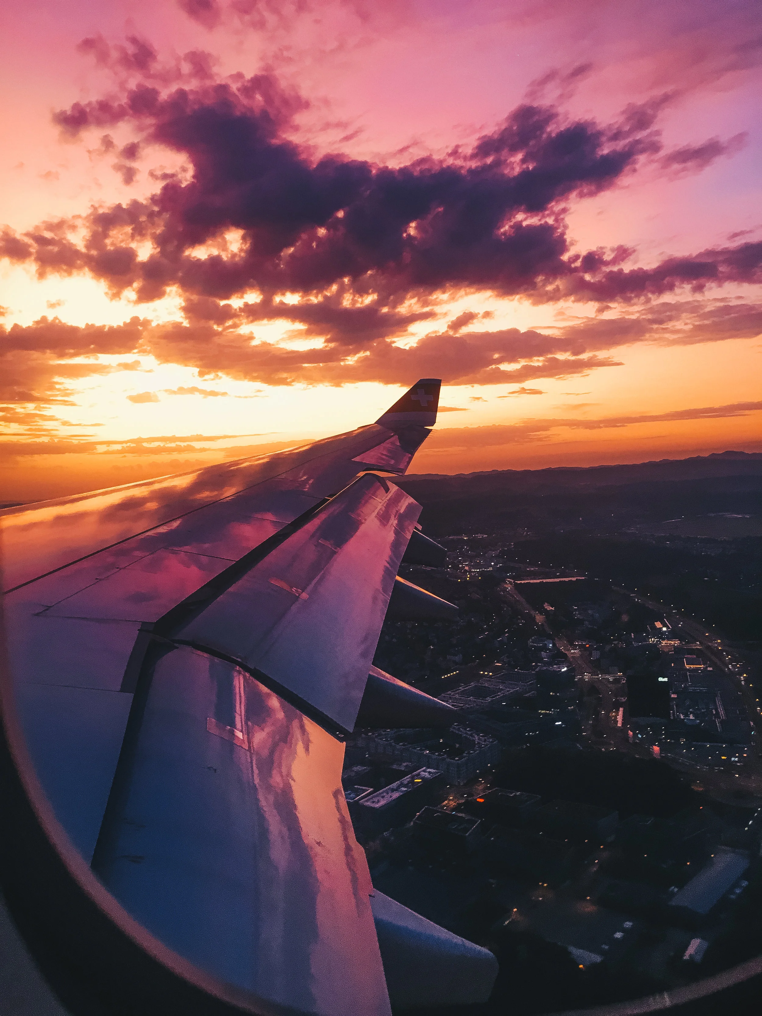 View of airplane wing during sunset with colorful sky and city lights below.
