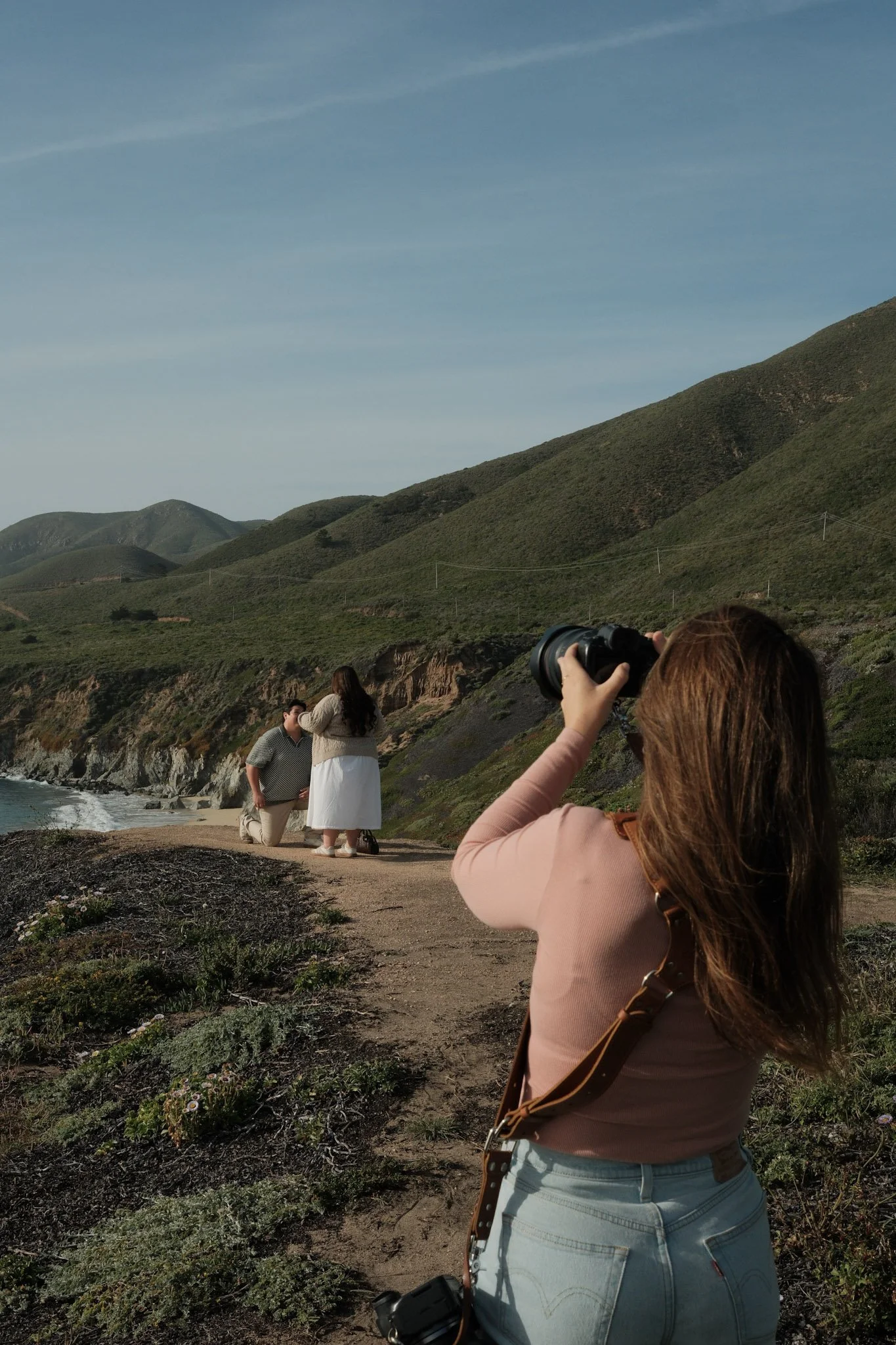 Big Sur Proposal Photographer taking a photo of a couple on a beach with green hills in the background. The woman is  holding a camera, with another camera hanging from her shoulder. The are on the cliffs of Garrapata in Big Sur, Ca