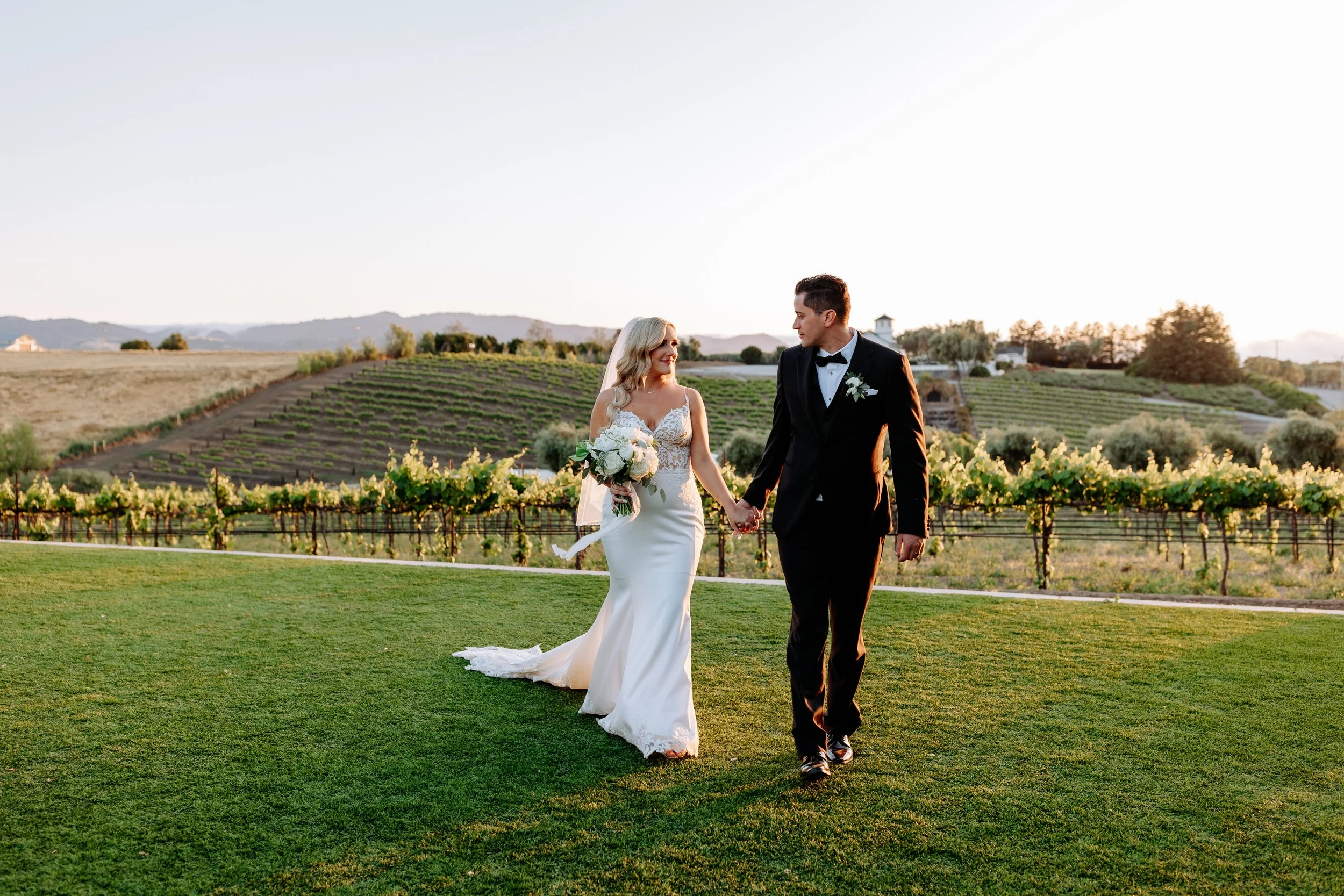 A bride and groom walk hand in hand through a vineyard during sunset, with rolling hills in the background at Leal Vineyards wedding.