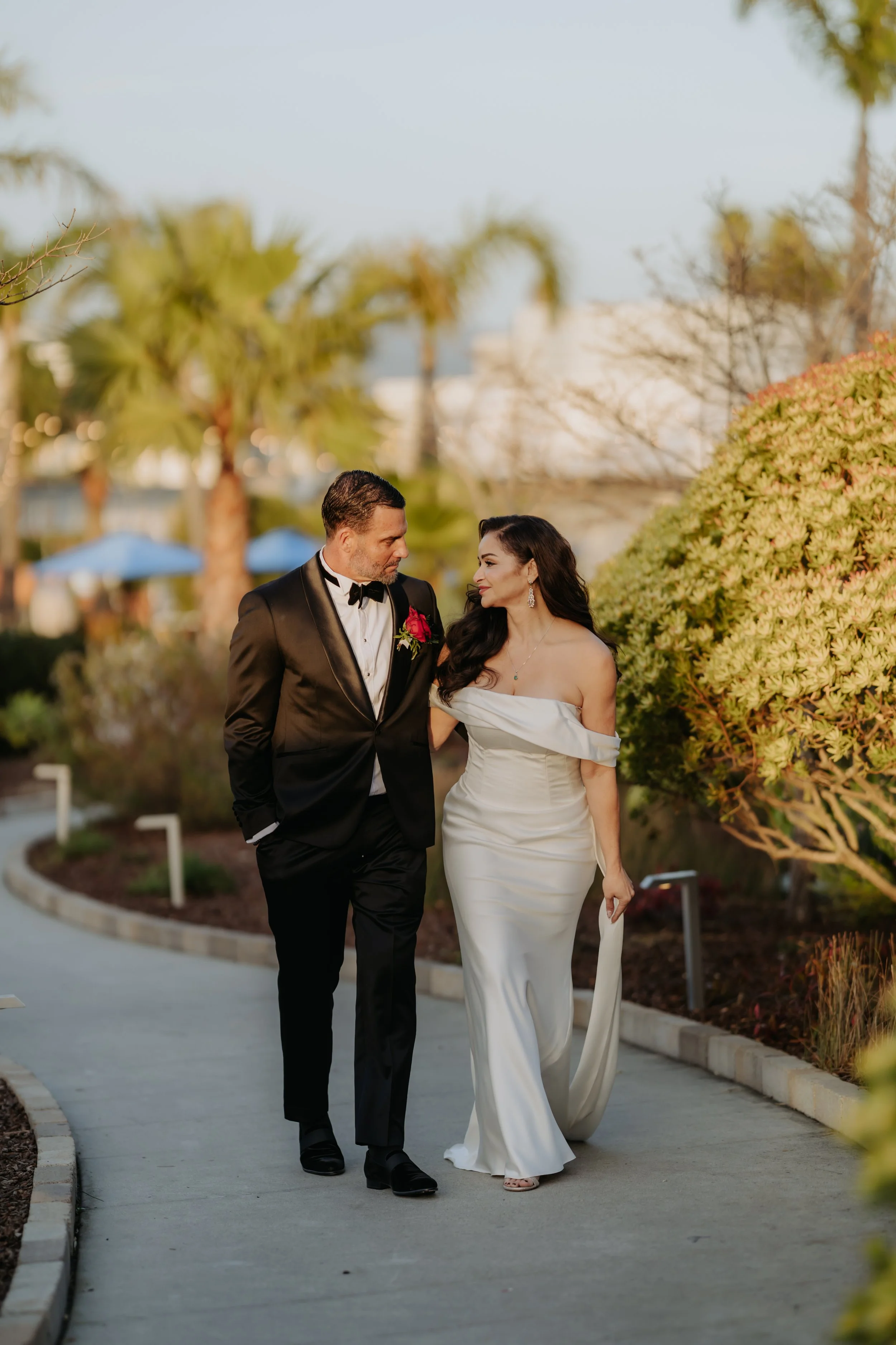 A couple dressed in formal wedding attire walking outdoors in a park or garden with palm trees and bushes, during daylight at The Dream Inn Santa Cruz, Ca.