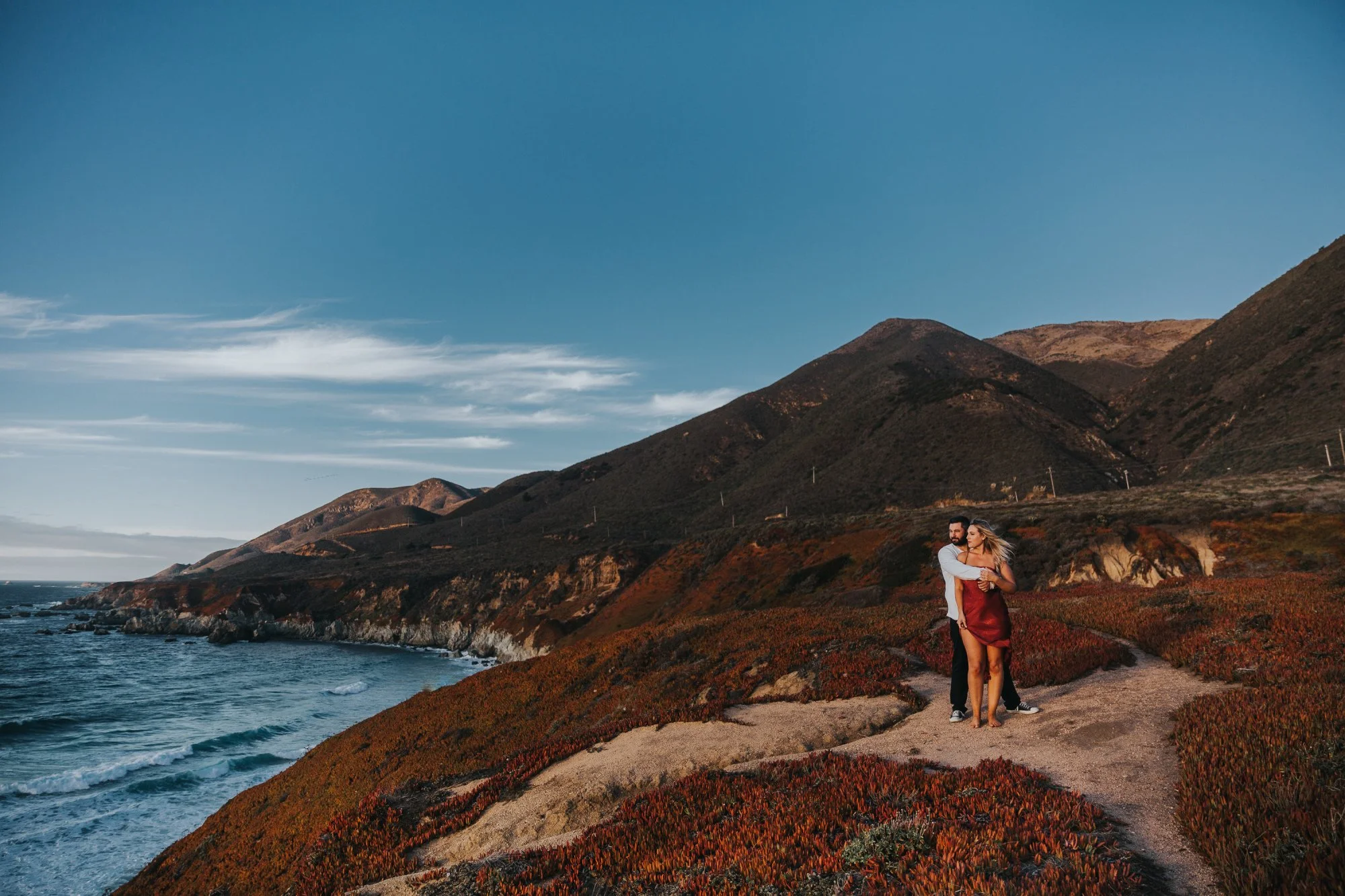 A couple embracing along a coastal trail with mountains in the background and the ocean on the left, under a partly cloudy sky celebrating their engagement photos in Big Sur, Ca.
