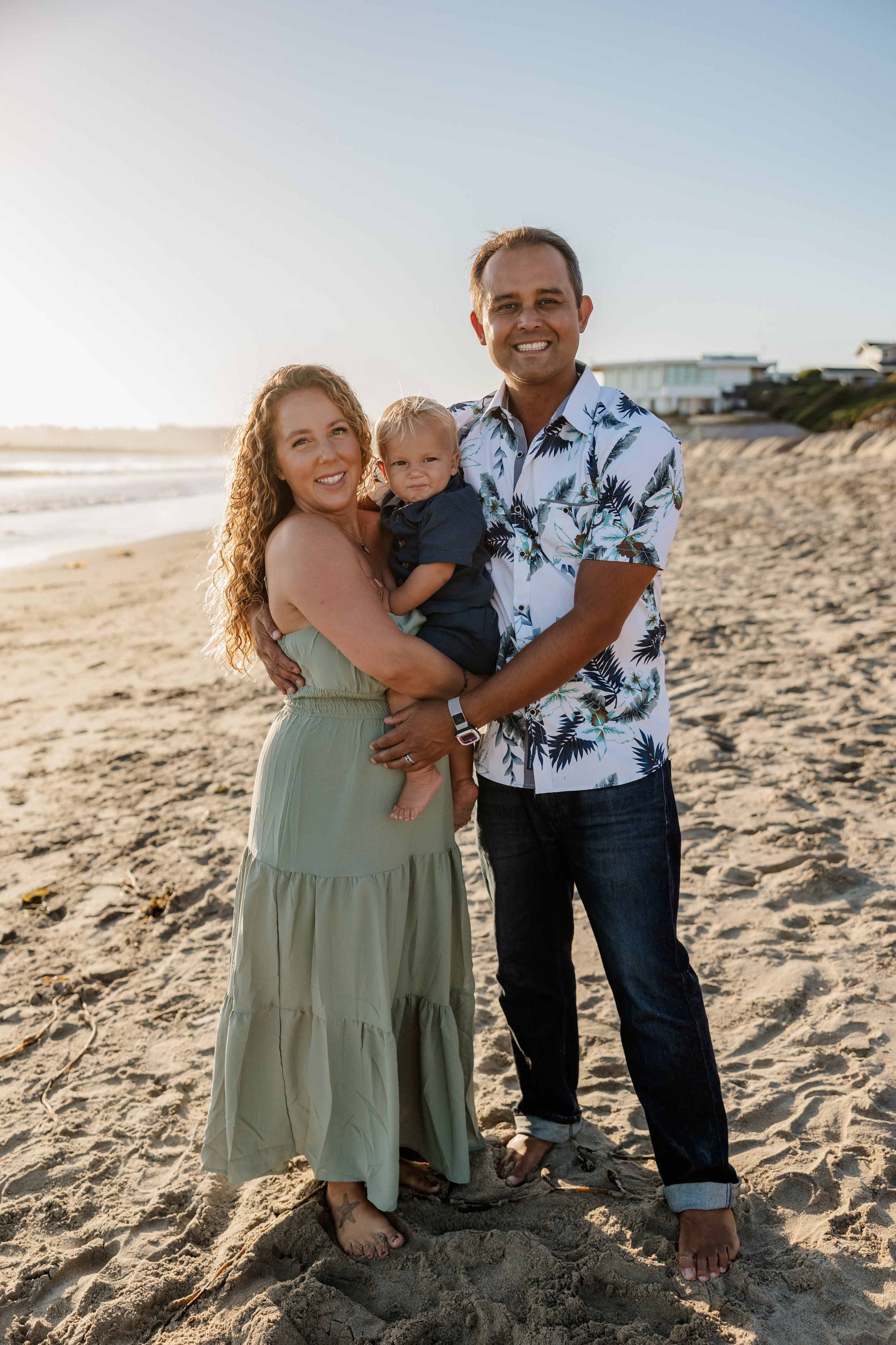 A happy family of three at the beach during sunset, with the woman holding a young child and the man standing beside them, all smiling in Santa Cruz, Ca.