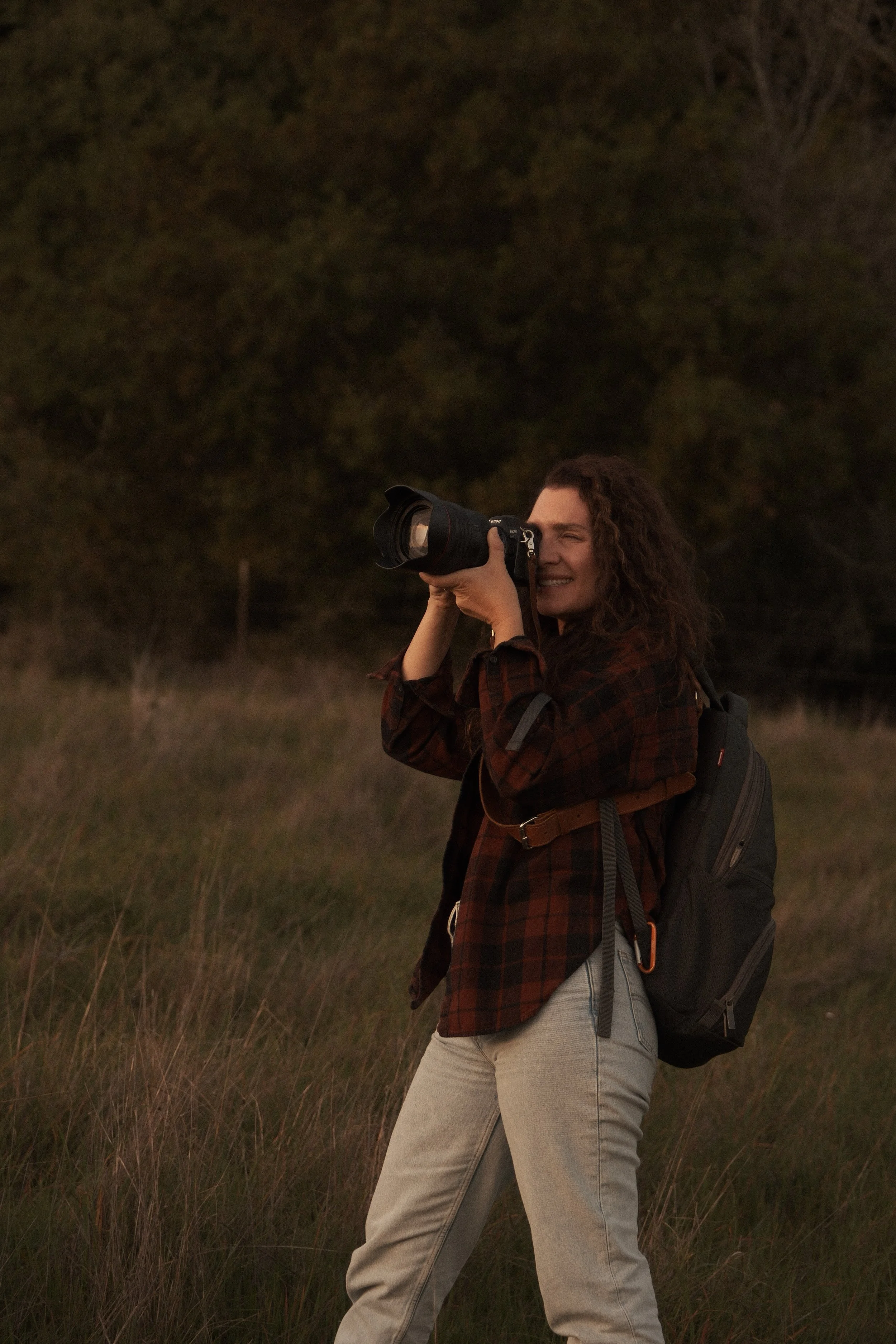 Kassandra Thomsen Santa Cruz Wedding Photographer standing outdoors in a field with tall grass, holding a DSLR camera up to her eye, smiling, with a backpack on her back, during sunset or dusk.
