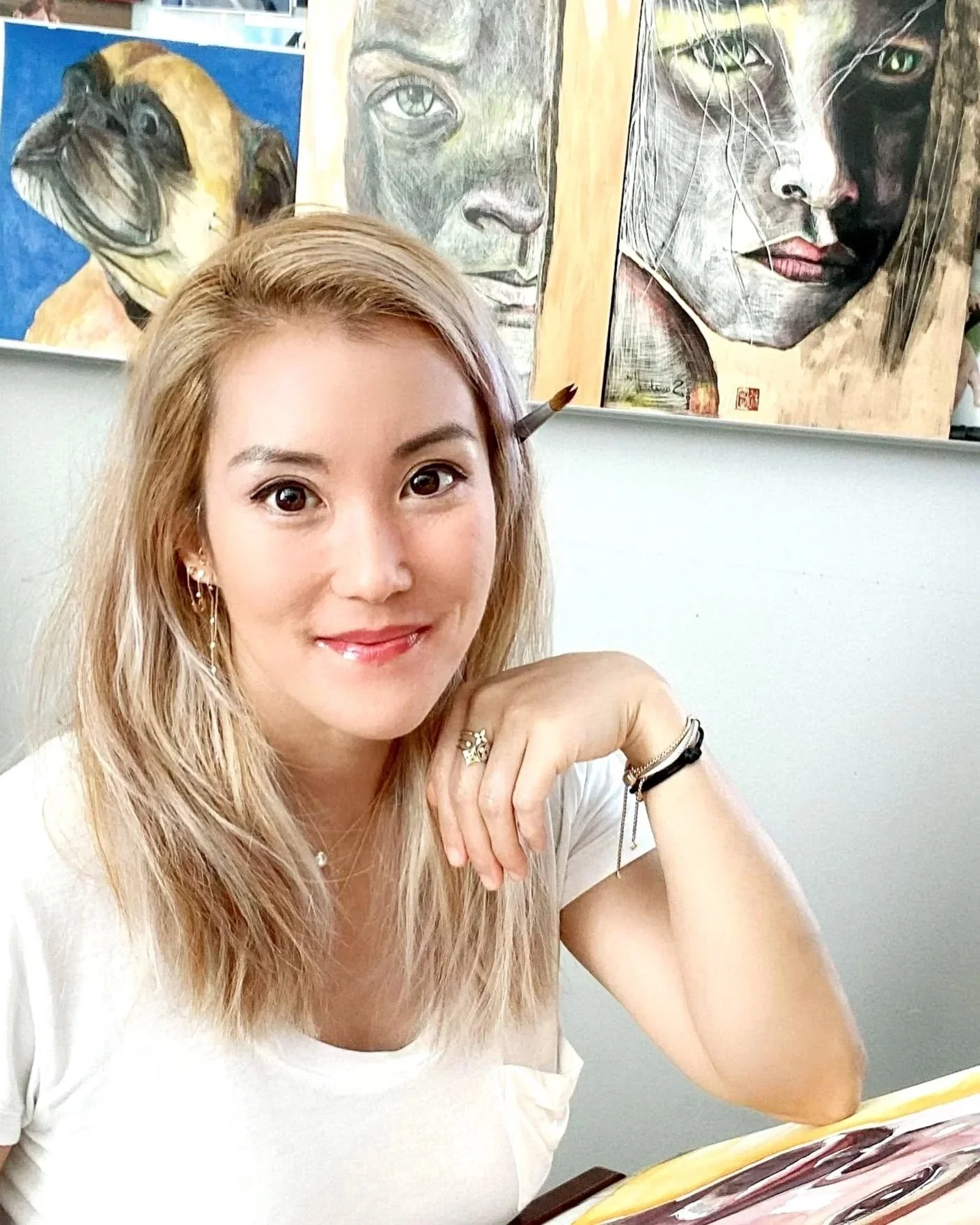 A young woman with long blonde hair and jewelry is smiling at the camera while sitting at a table. Behind her are three paintings of human faces and a dog.