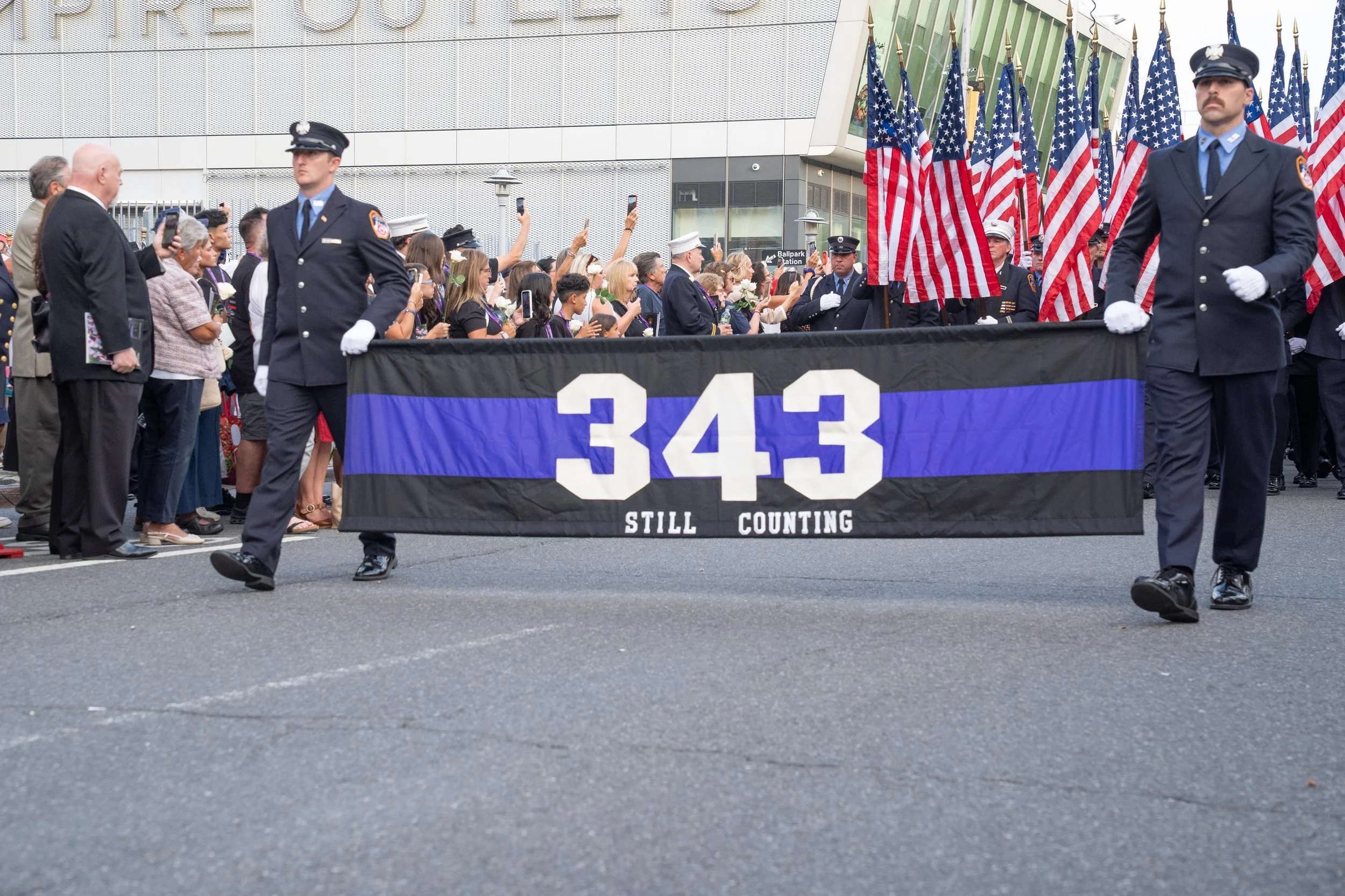 FDNY members carry a banner reading “343 Still Counting” beside a ballpark as spectators watch a memorial procession.