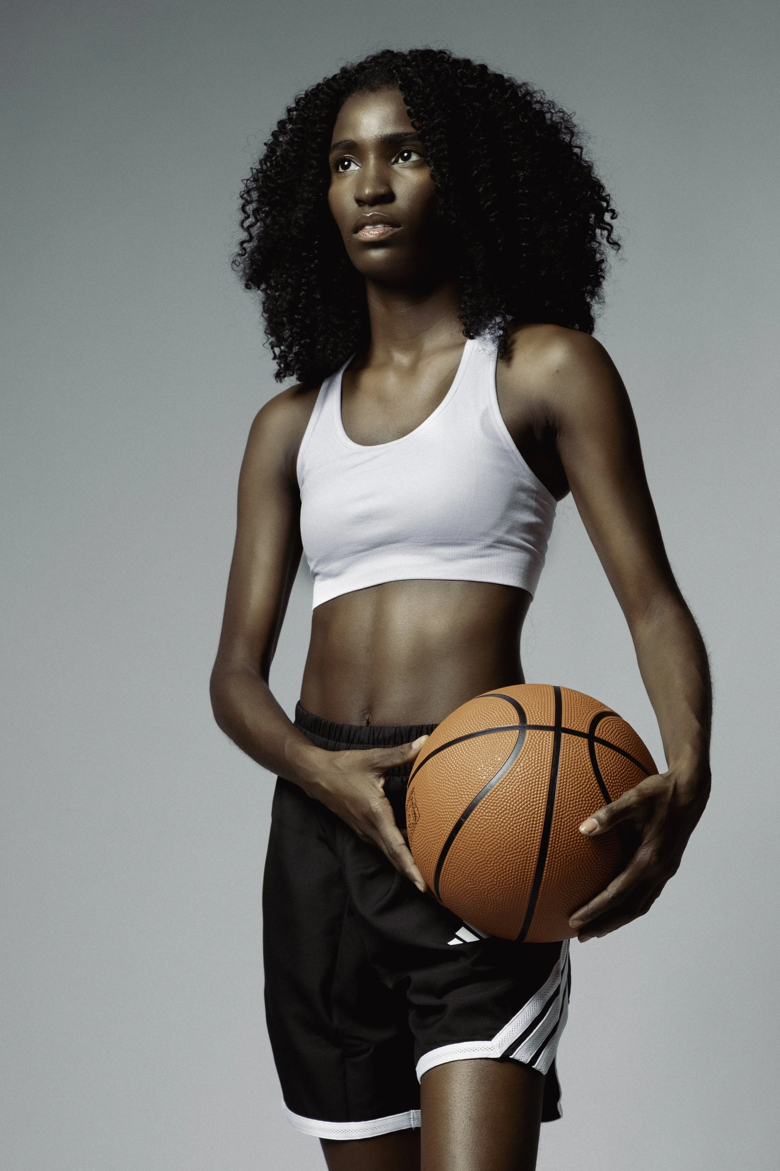 Athletic studio portrait of a woman holding an orange basketball, wearing a white crop top and black shorts against a gray background.