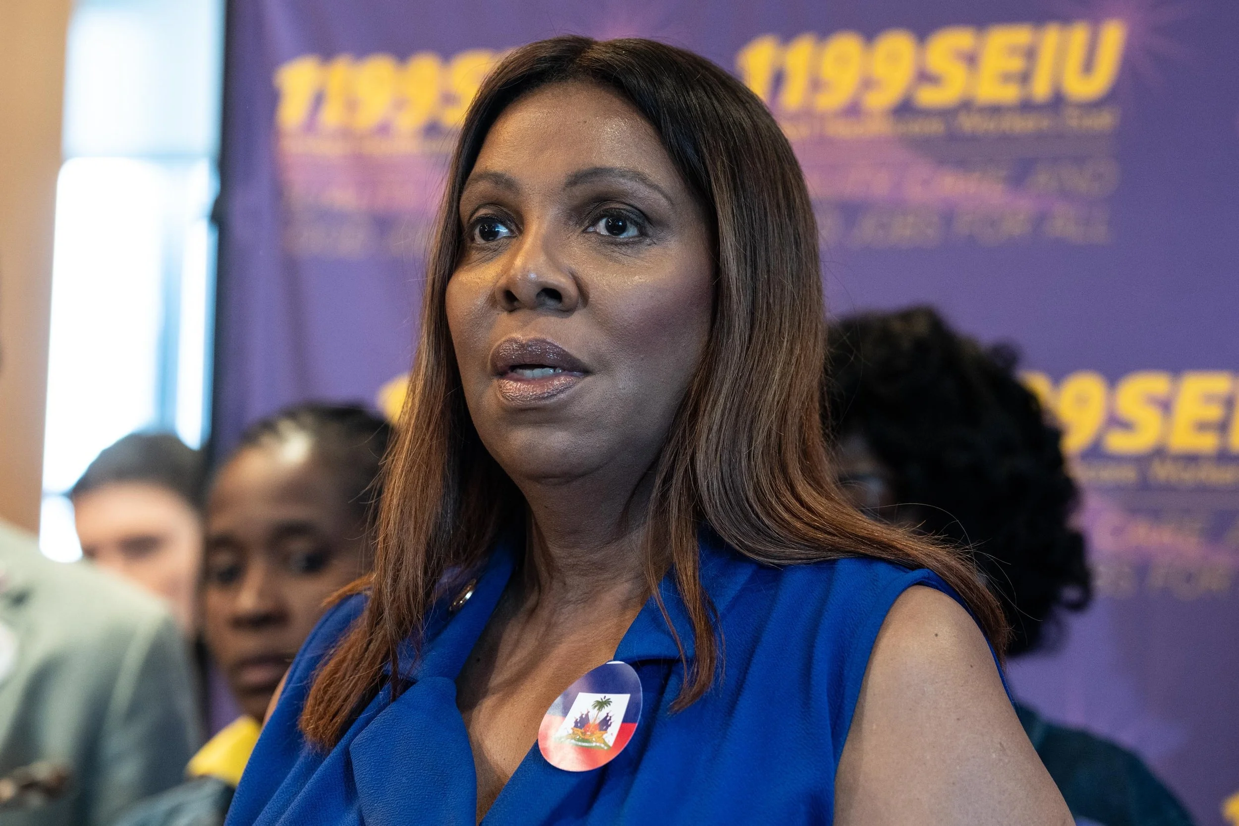 A speaker in blue stands in front of a purple 1199SEIU backdrop, wearing a button depicting the Haitian flag.