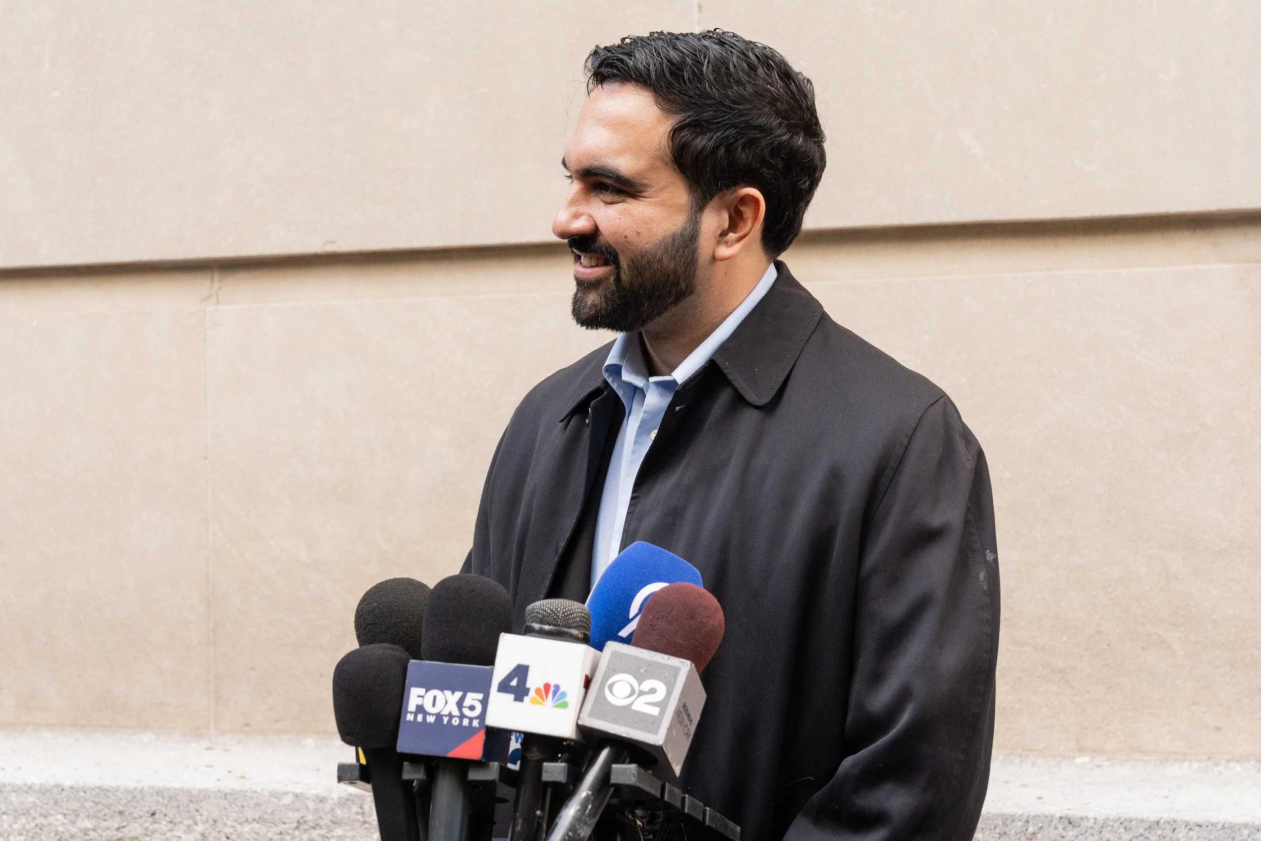 A man speaks to a cluster of television news microphones outside a stone building.