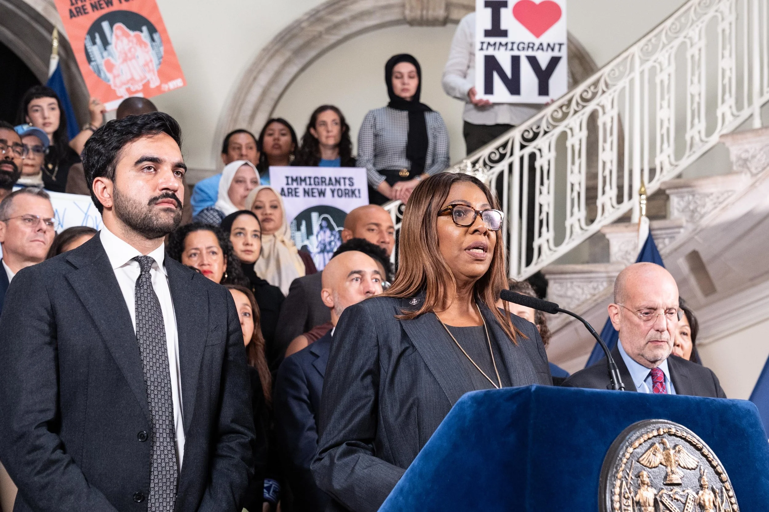 A speaker addresses a City Hall press conference with immigrant advocacy signs displayed behind the podium.