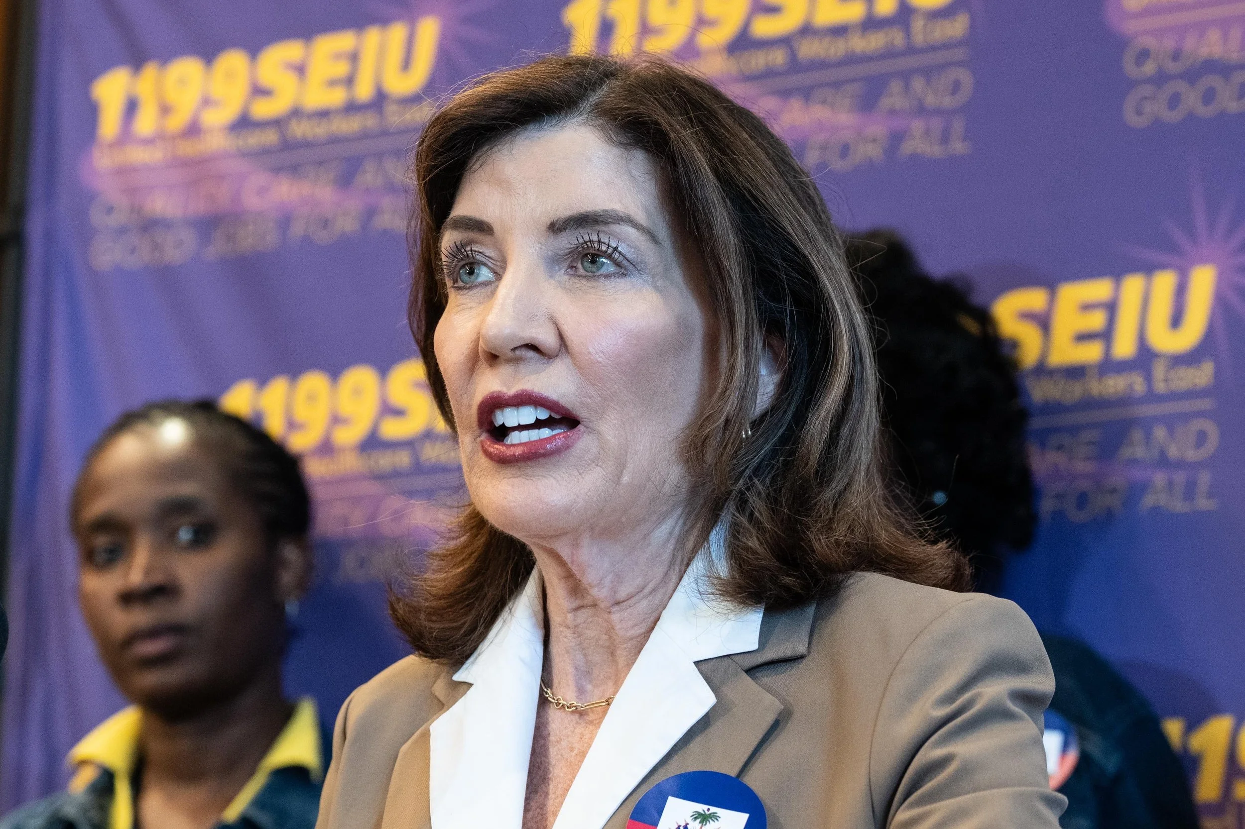 A speaker in a tan jacket addresses an event in front of a purple 1199SEIU backdrop.