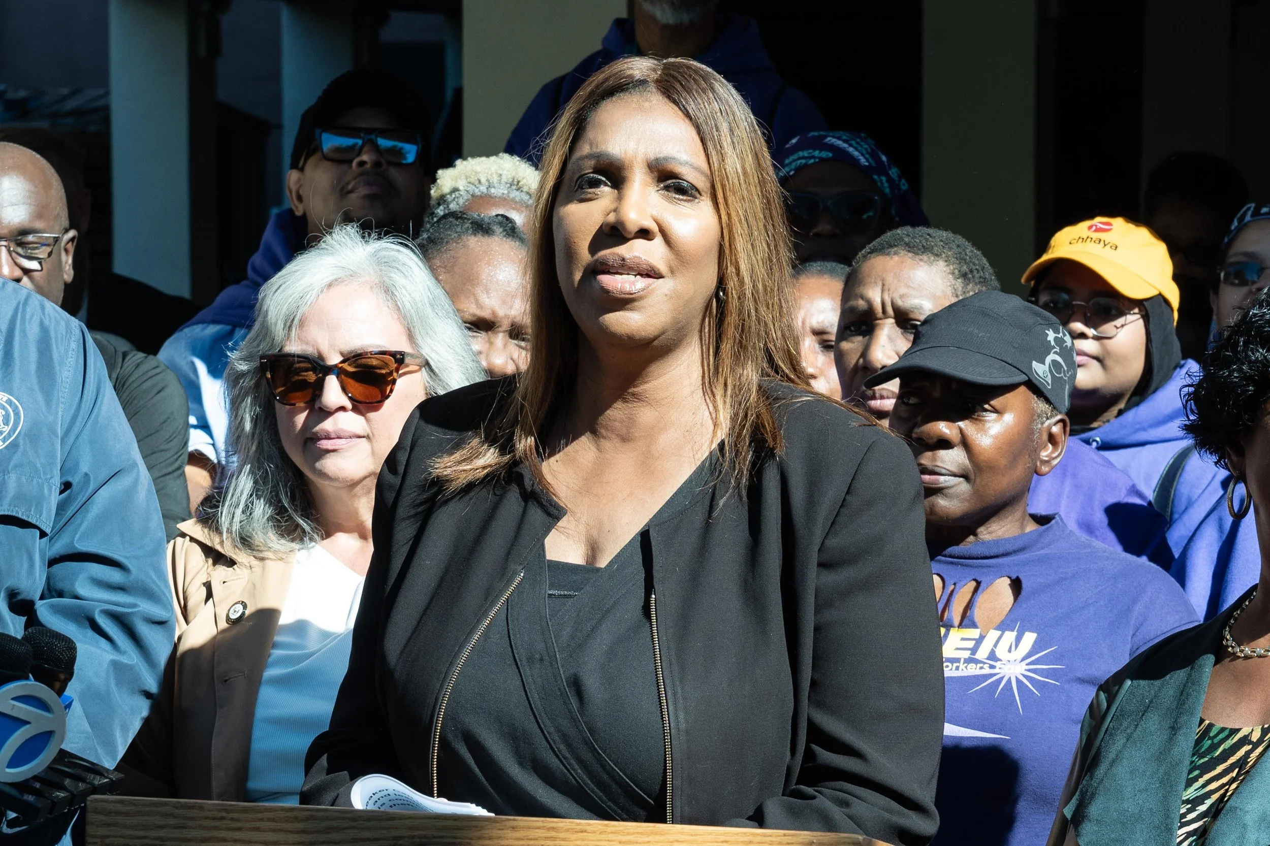 A speaker stands at an outdoor press conference with supporters wearing 1199SEIU clothing.