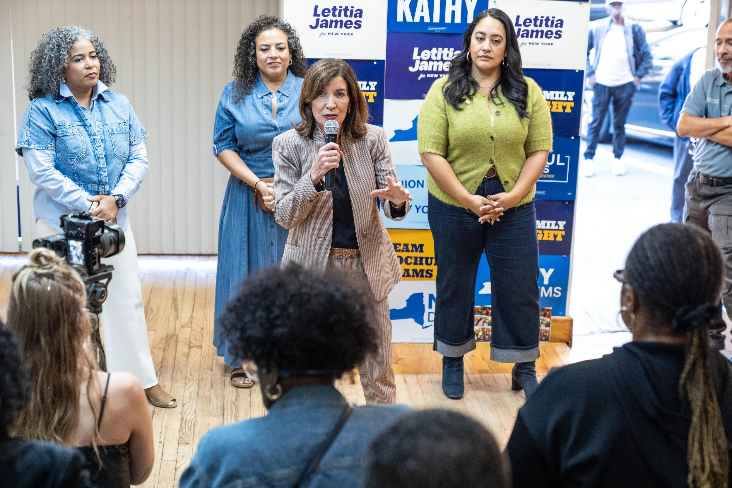 A speaker addresses a campaign gathering in front of signs for Kathy Hochul and Letitia James.