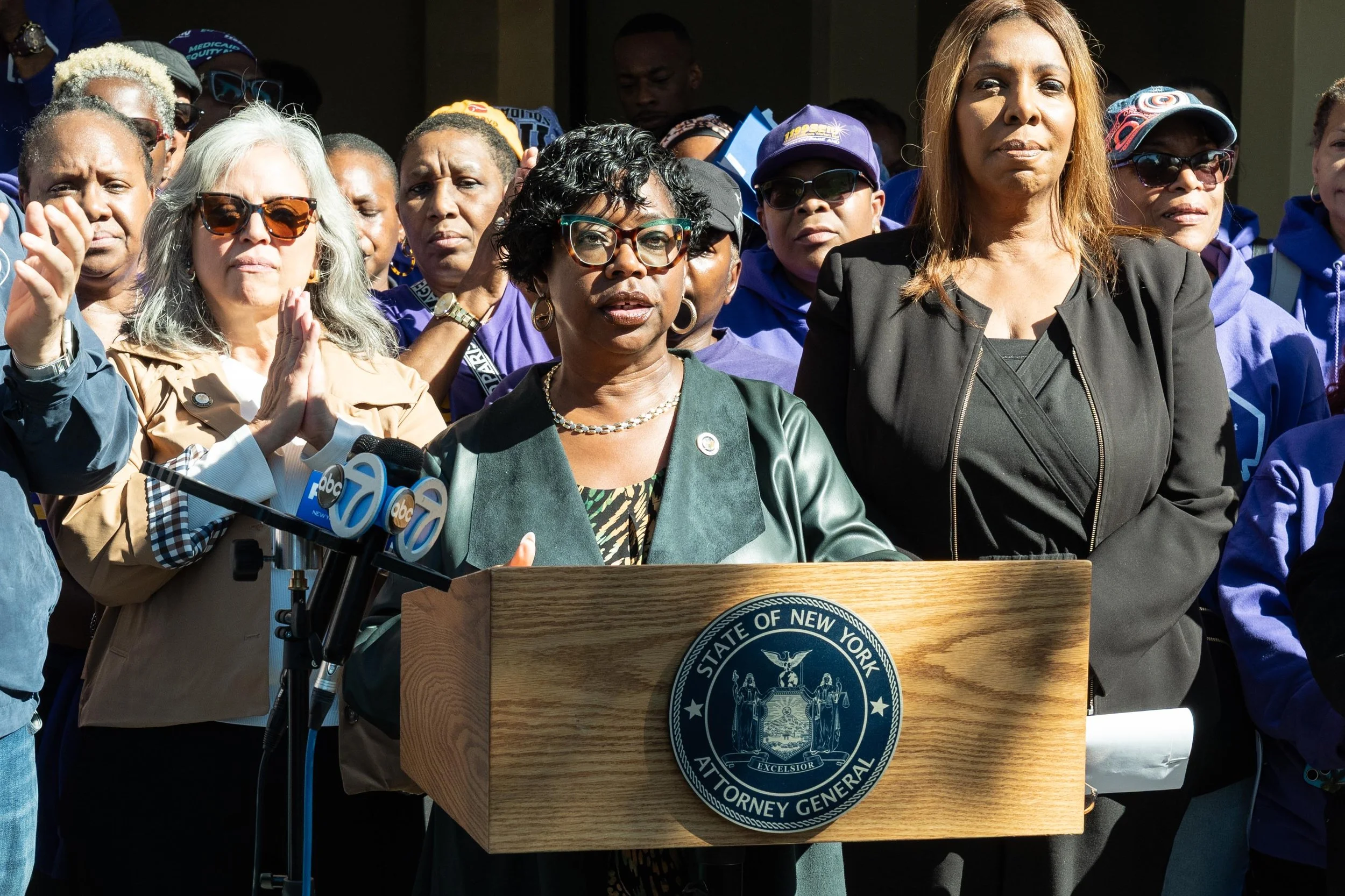 A speaker addresses reporters from a New York State Attorney General podium, with union members and community supporters behind her.