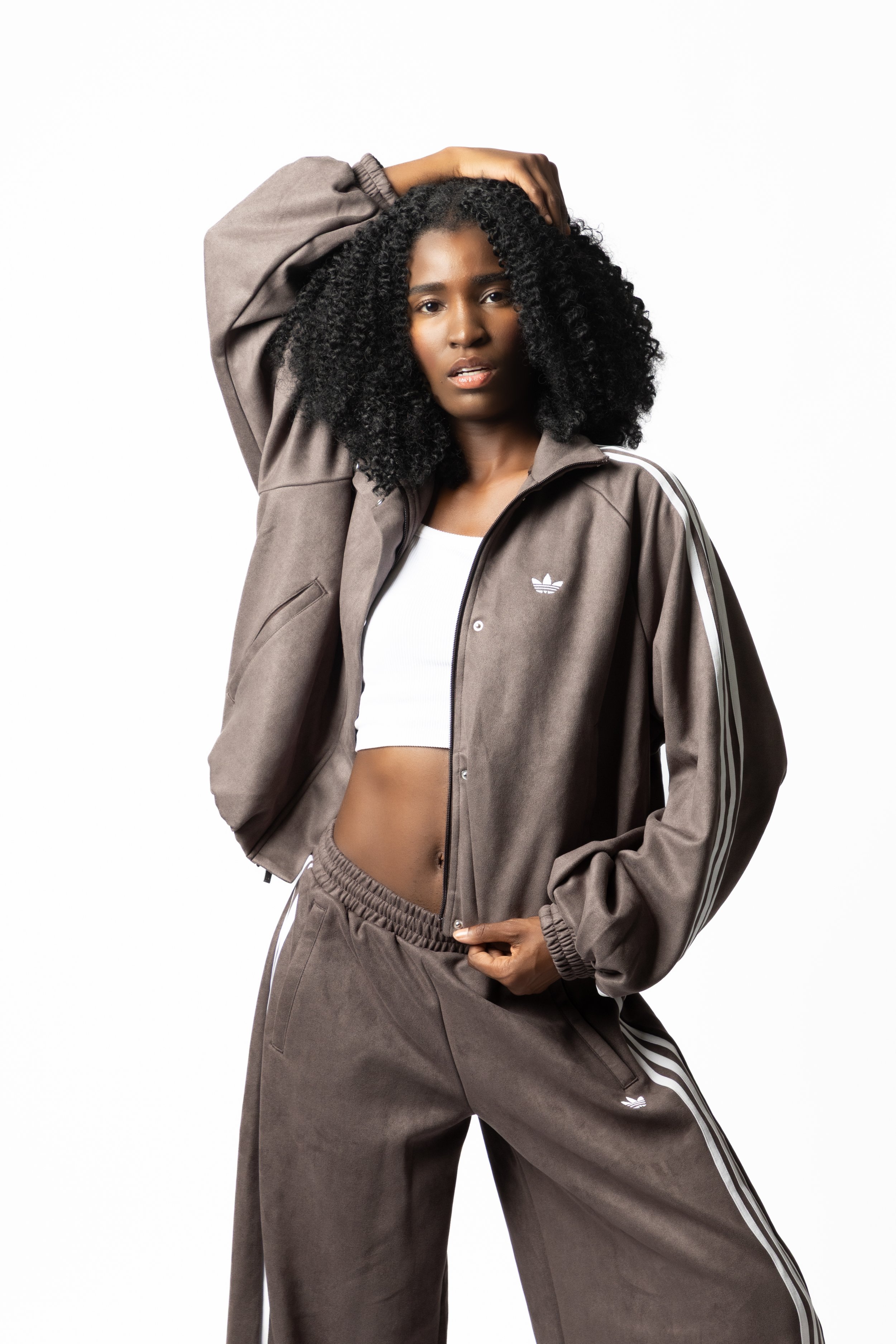 Fashion and athletic portrait of a woman in a taupe tracksuit, posing with one arm overhead against a light studio background.