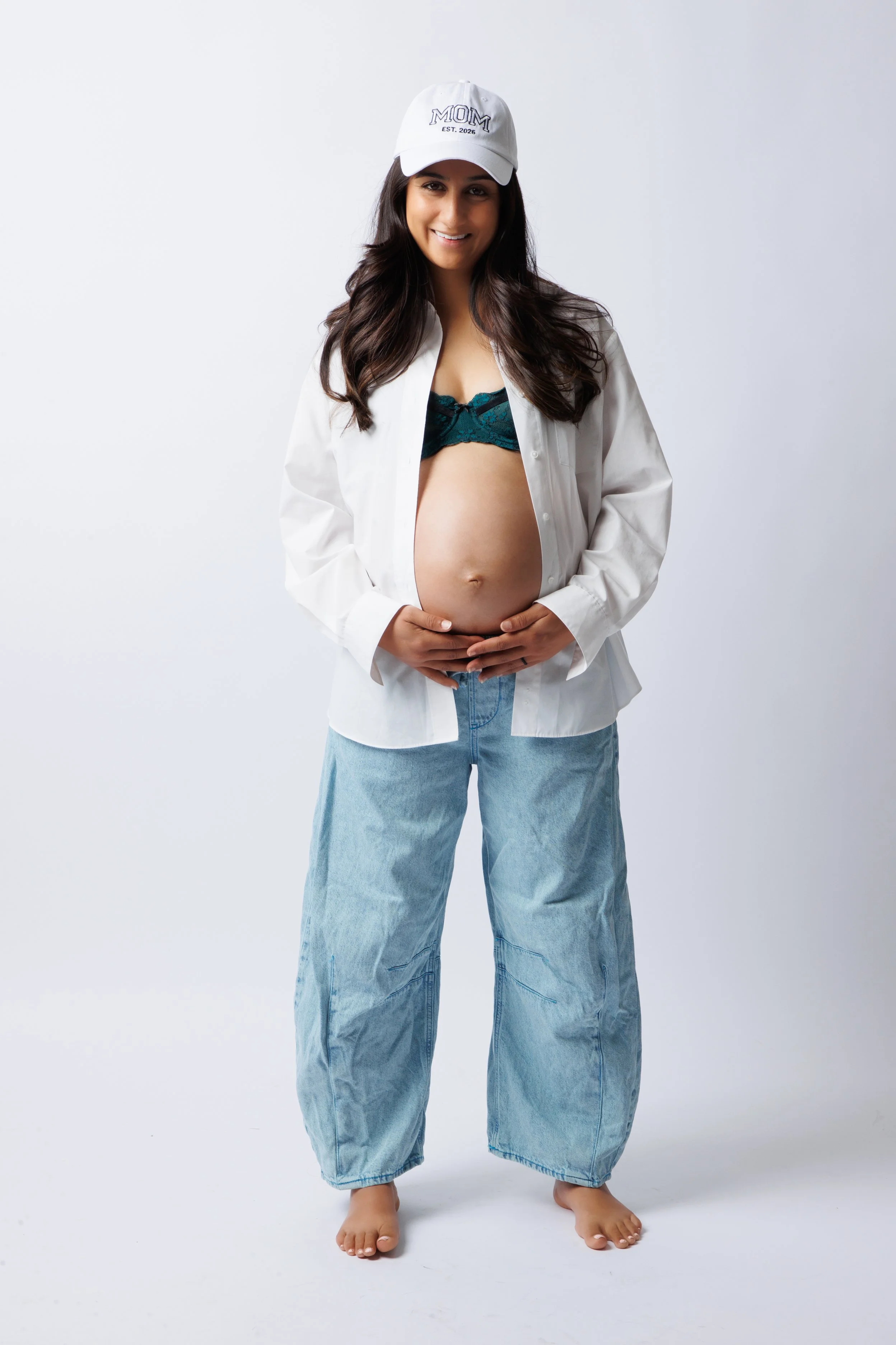 Full-length maternity studio portrait in a white shirt, jeans, and a white cap.