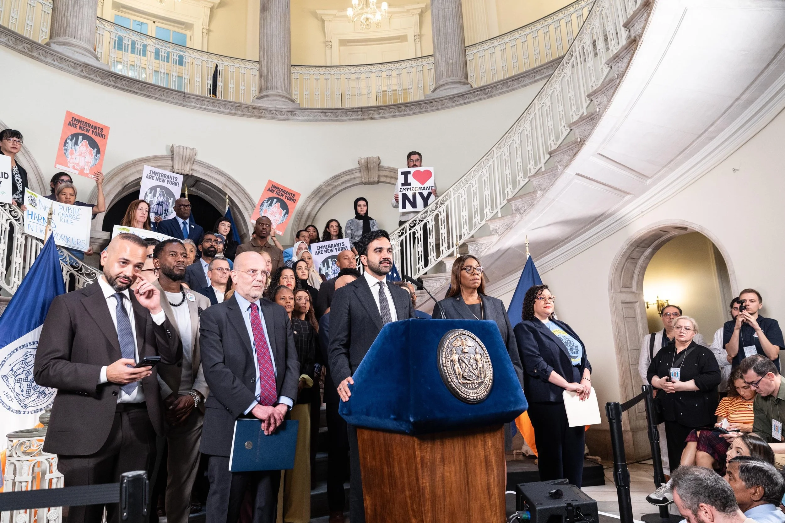 A wide view of a City Hall press conference, with speakers, supporters, immigrant advocacy signs, and members of the press in the rotunda.