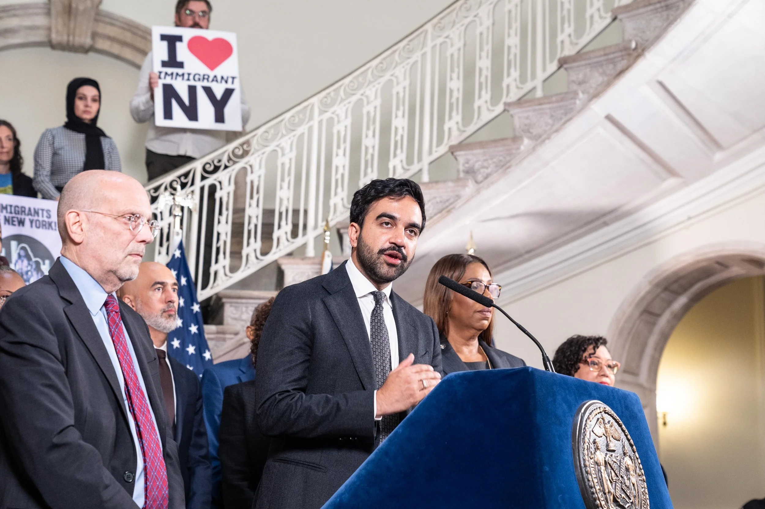 A speaker addresses reporters at a City Hall podium, with an “I Love Immigrant NY” sign in the background.
