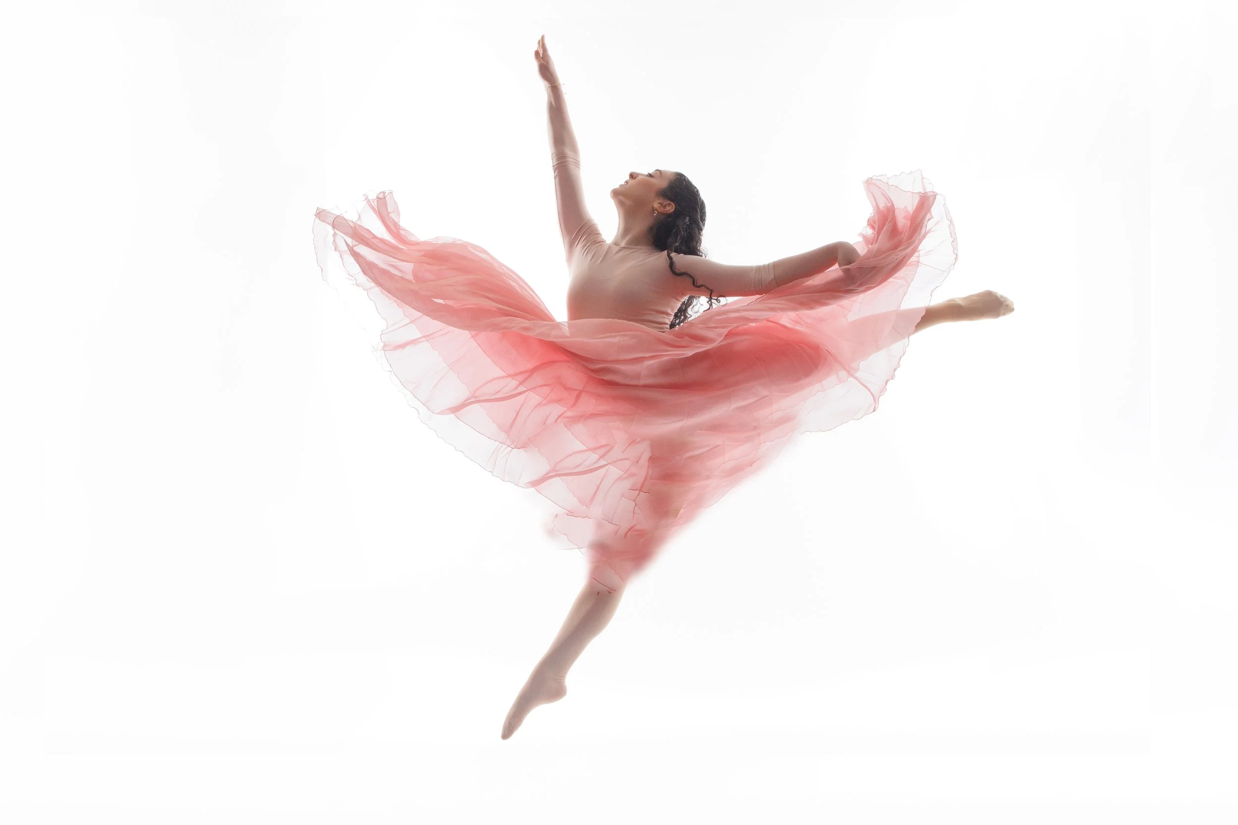 Dancer leaping in a flowing pink skirt with one arm extended overhead against a white studio background.