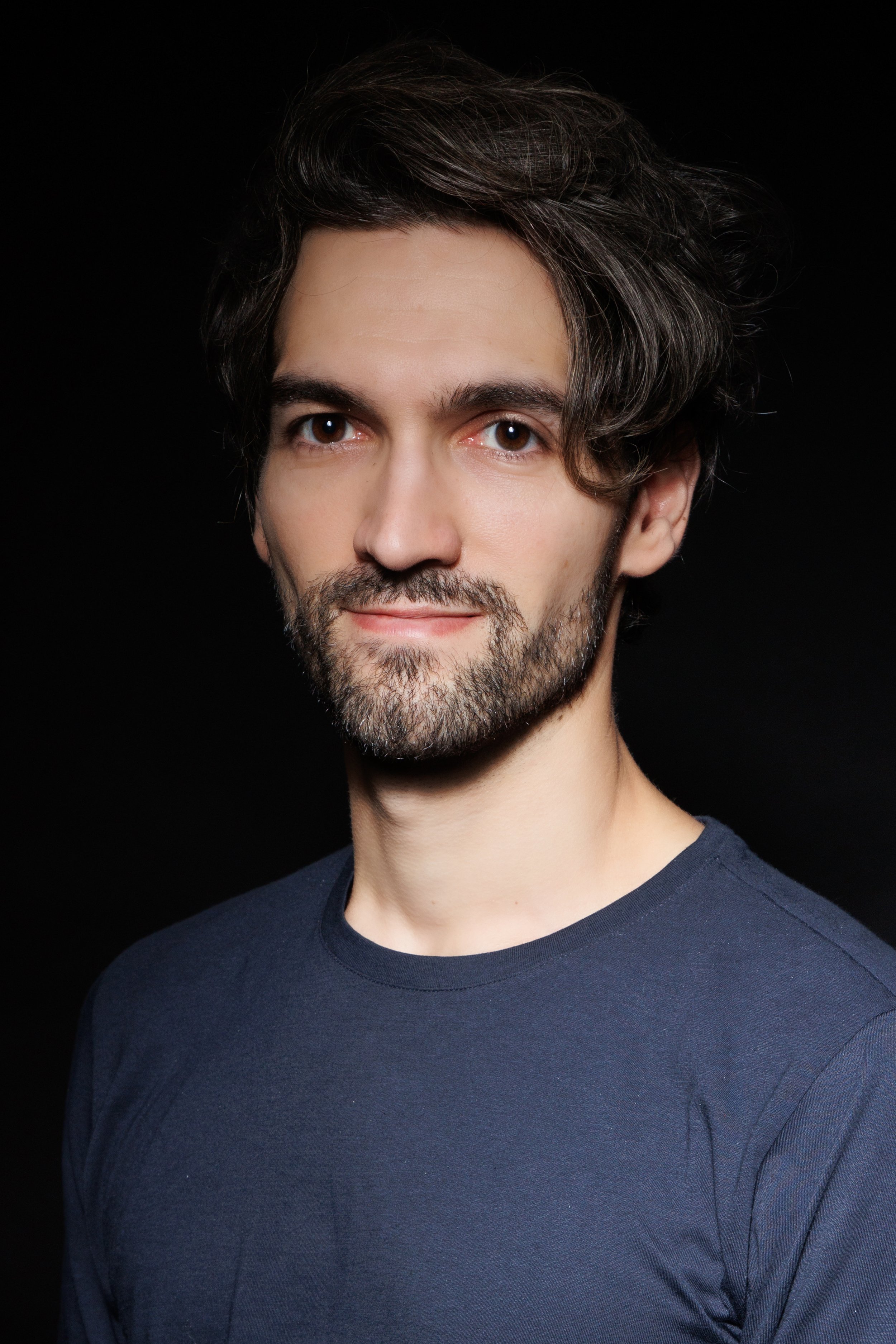 Studio headshot of a man with wavy hair and a beard wearing a navy shirt against a black background.