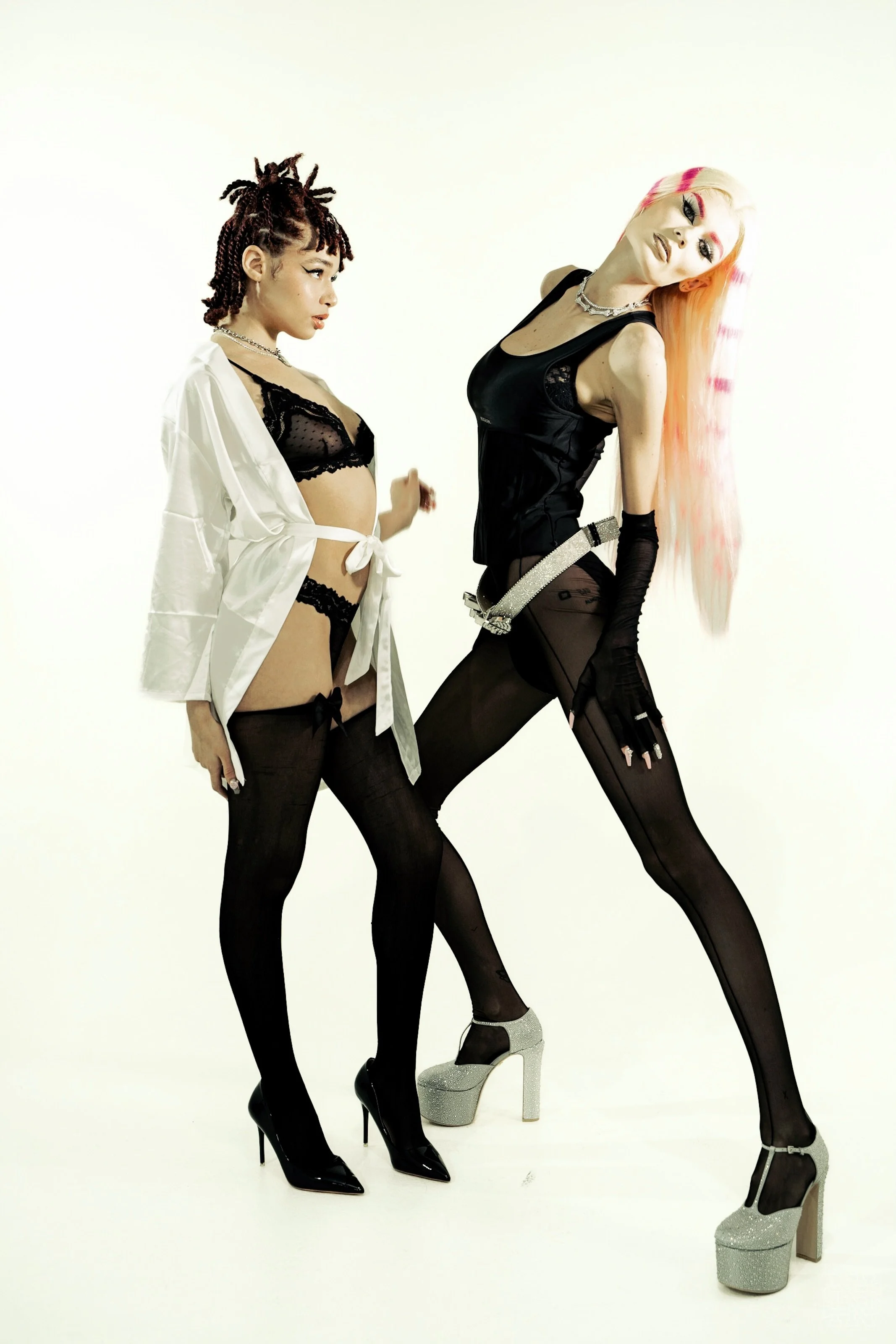 Two women in black lingerie and stockings with high heels, posing against a plain background. One woman has dreadlocks and a white robe partially open, the other has long, pink-tinted hair and wears a black satin top.