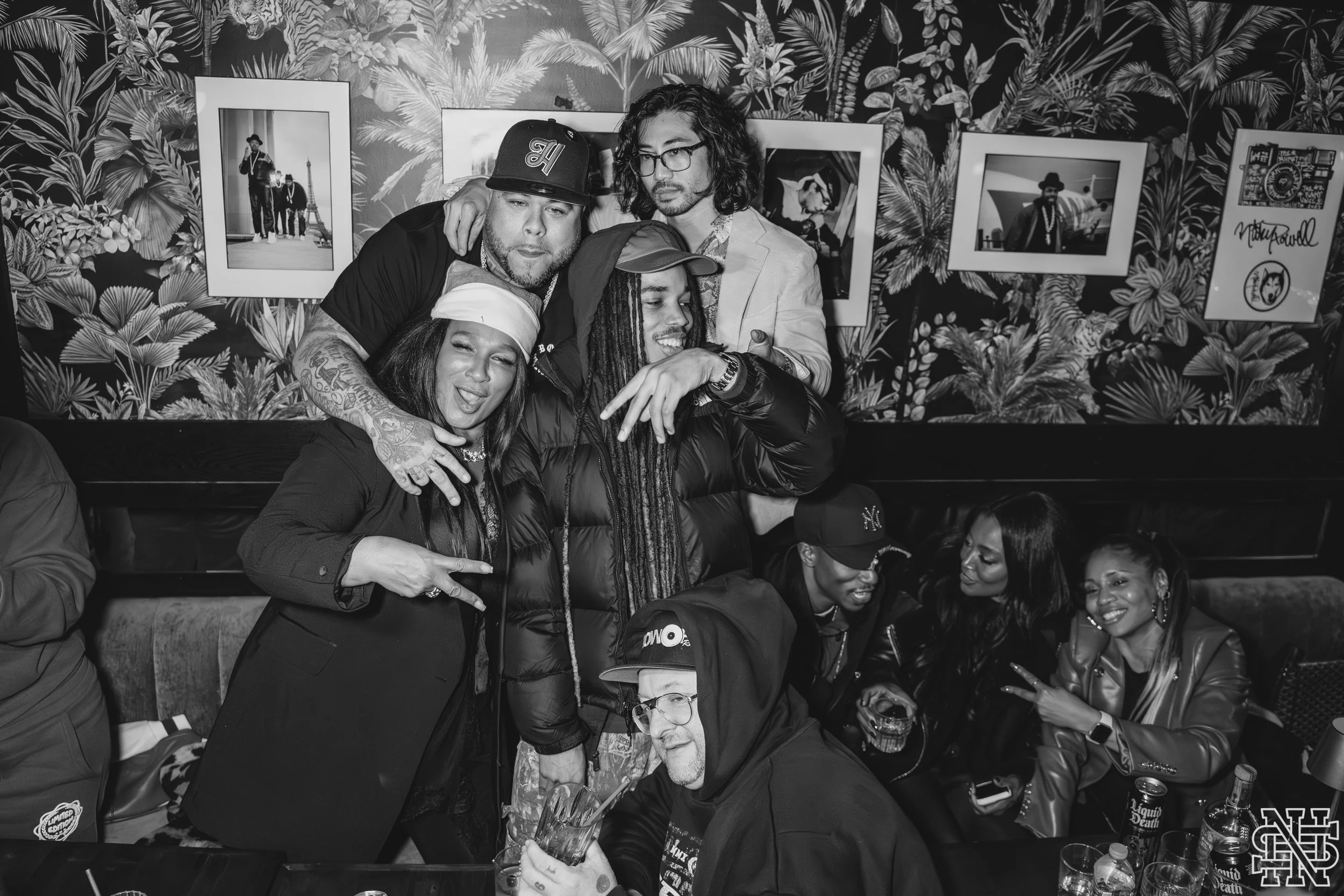 A group of eight diverse people celebrating together at a party, with some making peace signs and others smiling, in a room with tropical-themed wallpaper and framed photographs on the wall.