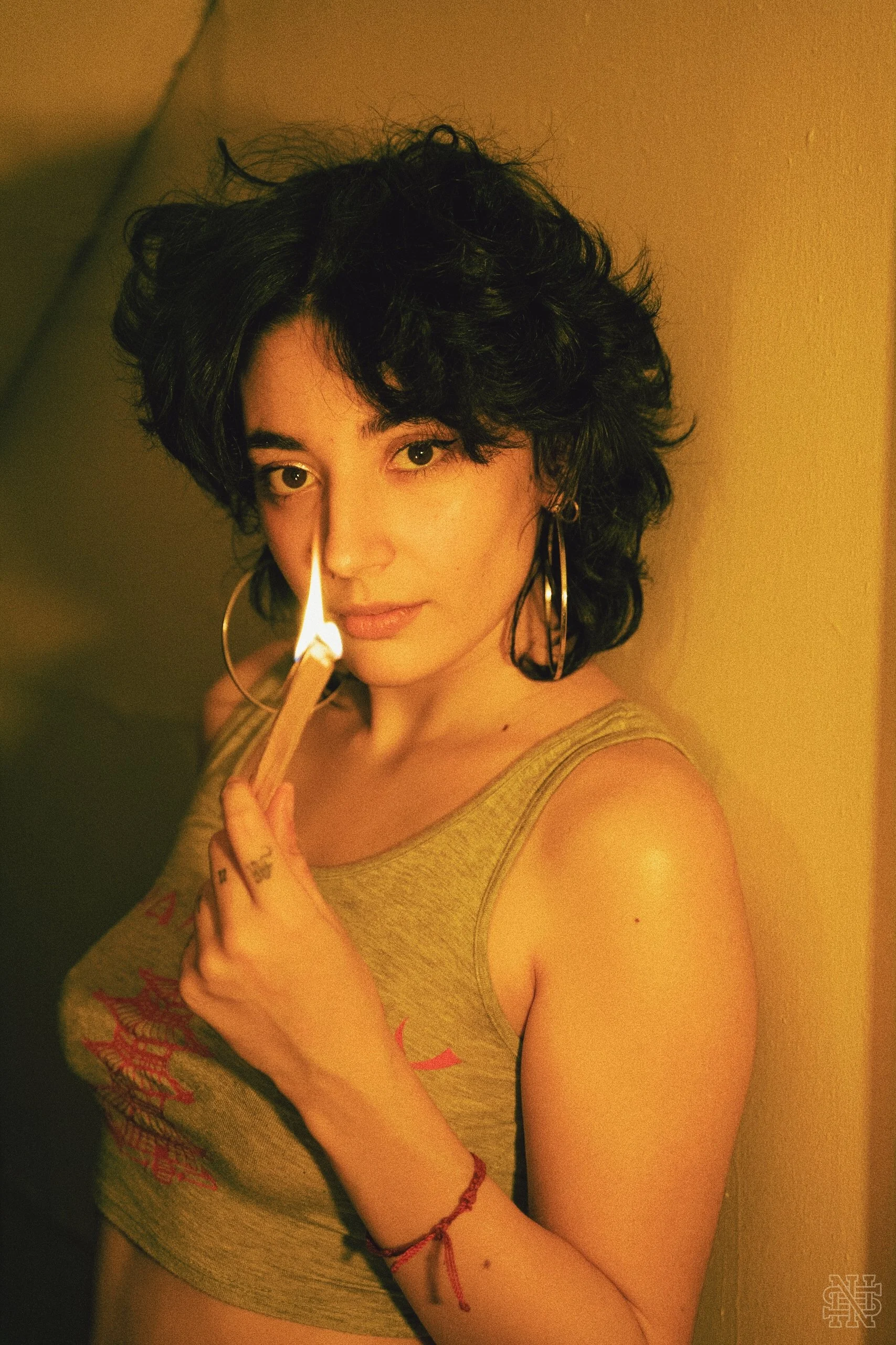 Young woman with curly dark hair and hoop earrings holding a lit match near her face against a yellow wall, wearing a sleeveless top and a red bracelet.