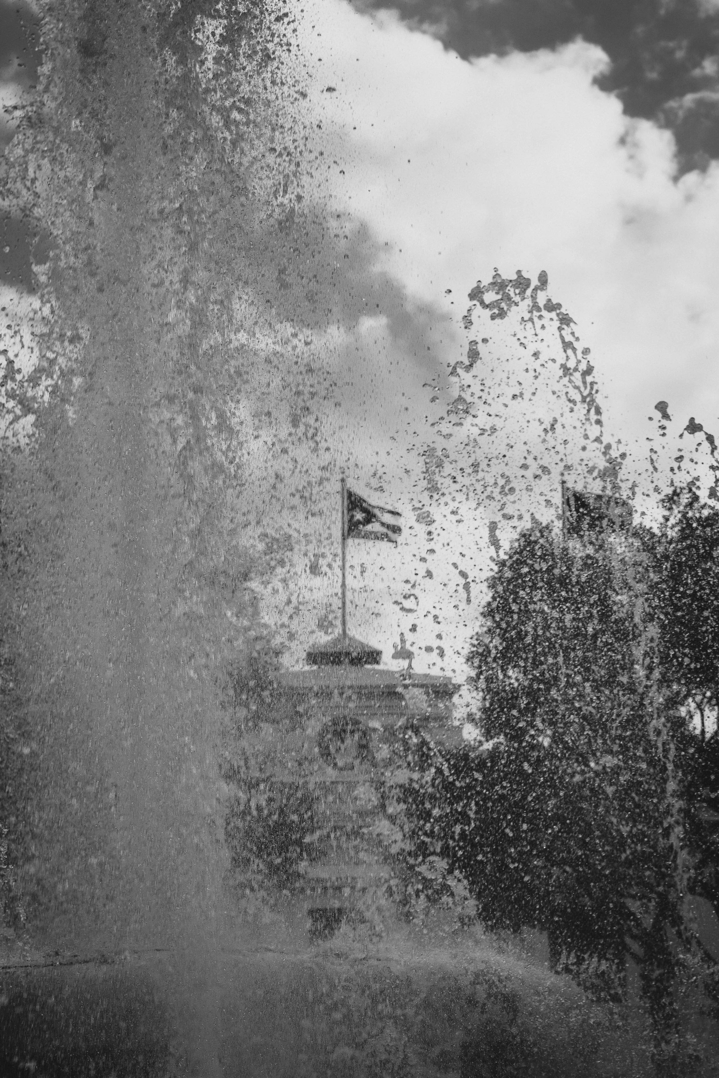 A splash of water in front of a U.S. flag on a flagpole, with trees and a cloudy sky in the background.