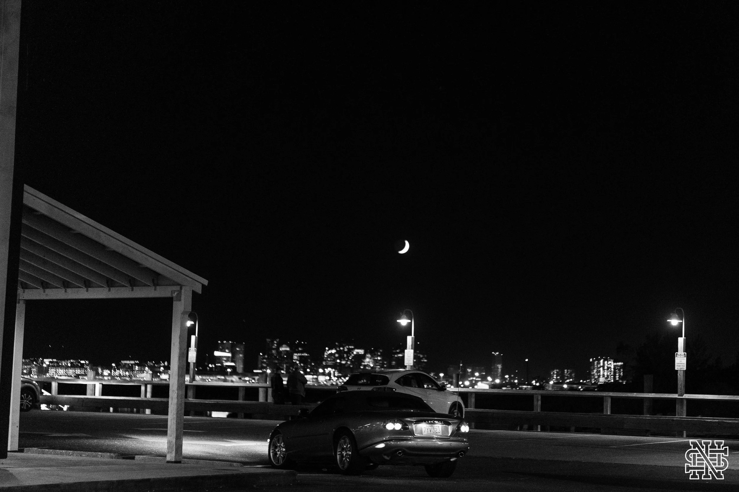 Night scene of a parking lot near a waterfront, with cars and a city skyline illuminated in the distance under a crescent moon.